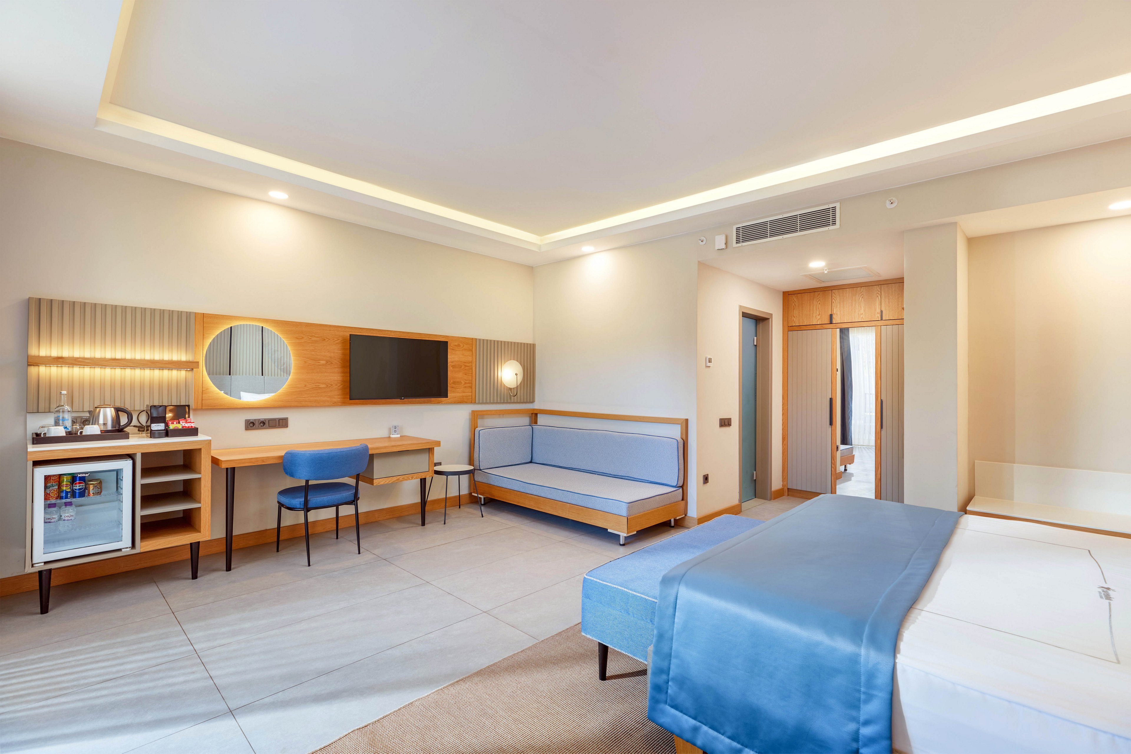 A spacious, contemporary hotel room features a neatly made bed with blue linens, a work desk with a chair, and a small sofa. The room includes a wall-mounted television, a round mirror, and a minibar with a visible refrigerator. Soft, neutral lighting and minimalist decor create a calm and inviting atmosphere.