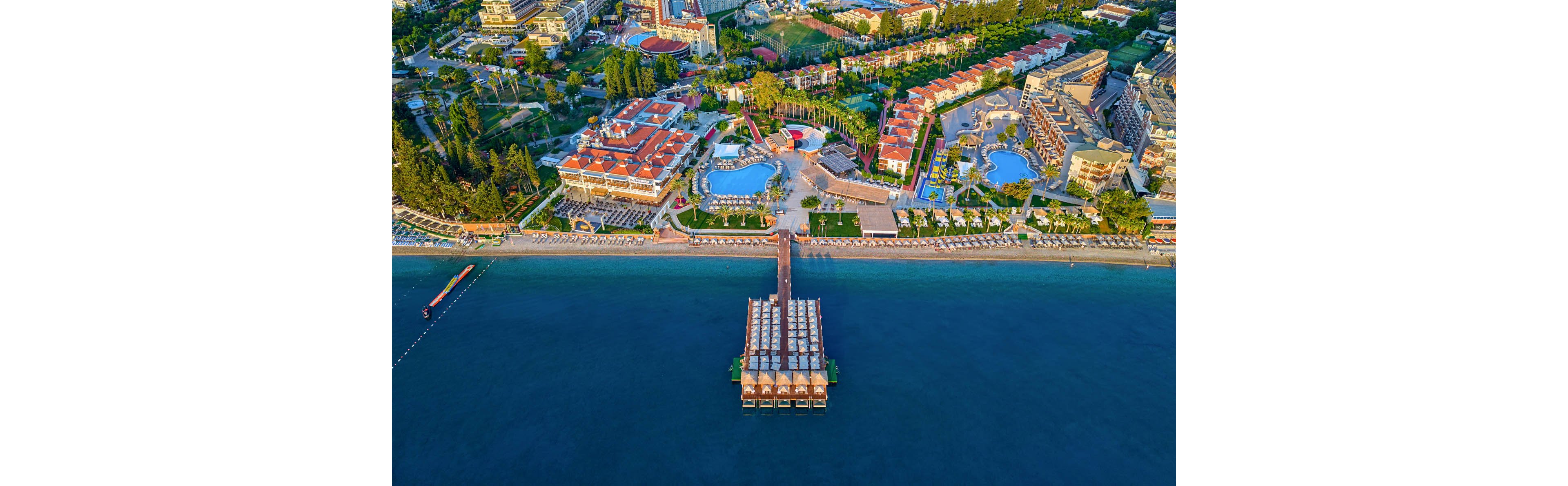 A large seaside resort complex is captured from above, showing multiple pools, buildings, and landscaped areas along the shoreline. A long wooden pier with covered seating structures extends into the calm blue sea. The scene highlights organized rows of beach chairs, pathways, and recreational facilities within the resort. Bright colors and clear weather create a vibrant, inviting vacation atmosphere.