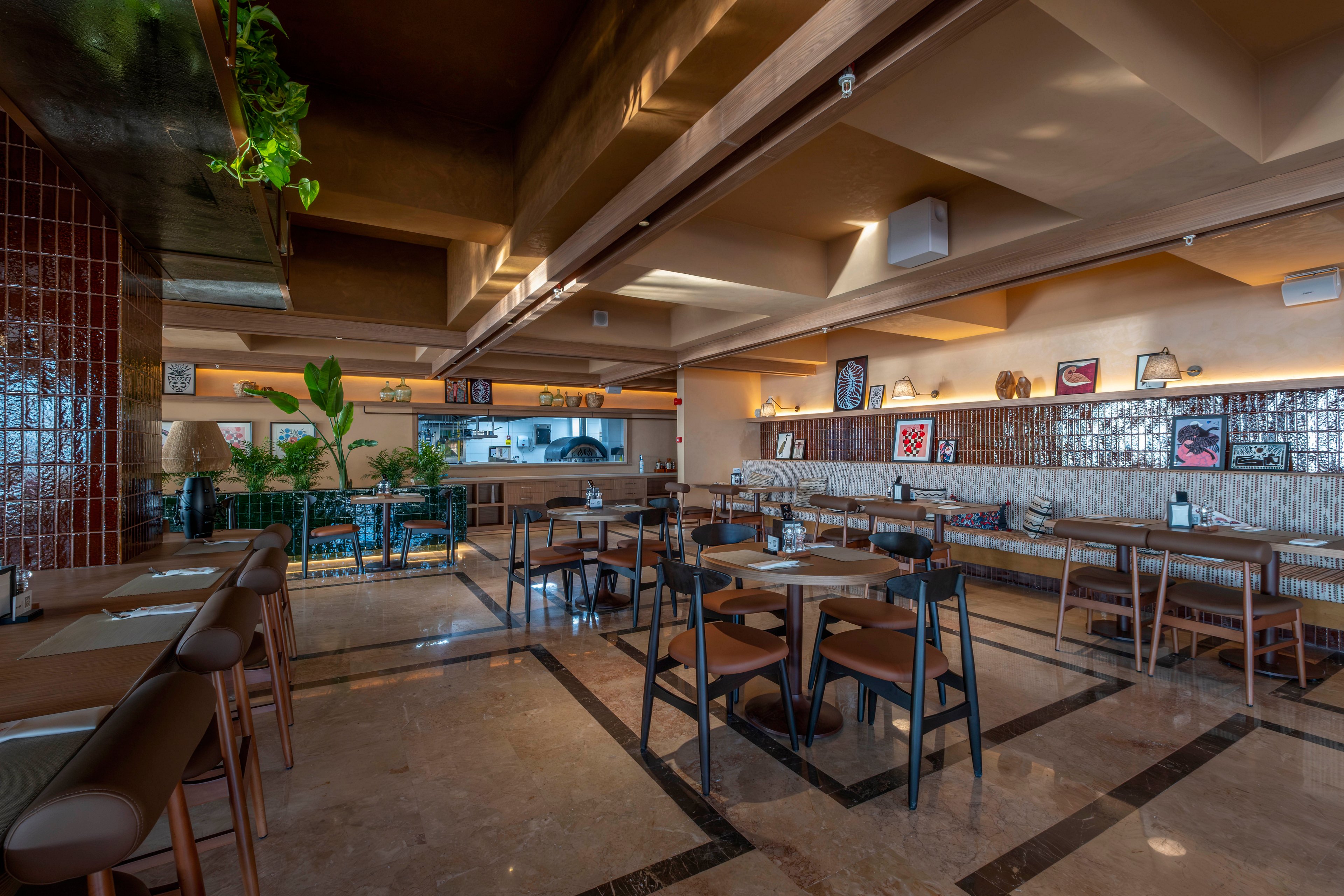 A contemporary restaurant interior features neatly arranged wooden tables and chairs in a spacious dining area. Warm ambient lighting highlights the beige and brown color palette, polished floor, and tiled wall accents. Green potted plants add freshness near the open kitchen and bar counter at the back. Framed artwork and decorative shelves create a cozy, stylish atmosphere in the otherwise empty space.
