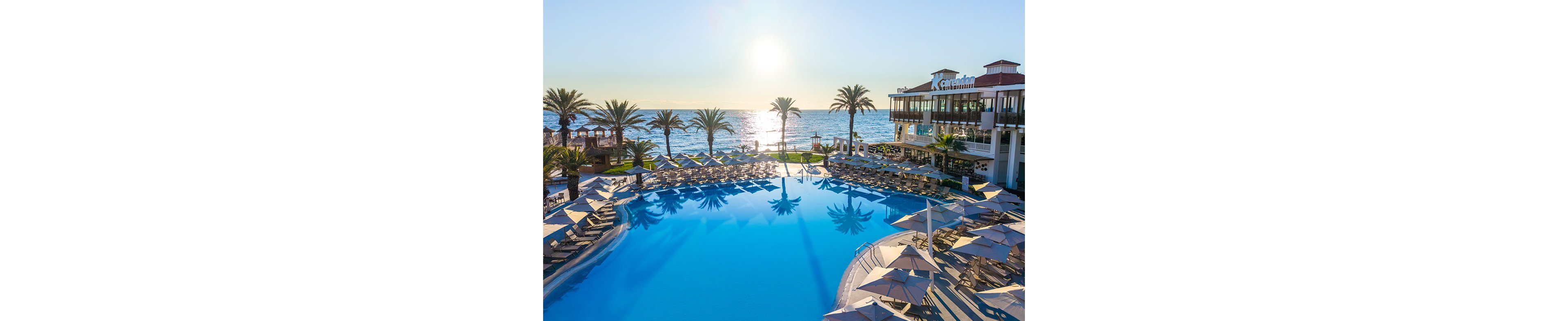 A large resort swimming pool reflects the bright sun setting over the sea. Rows of sun loungers and umbrellas surround the pool, creating a relaxing holiday atmosphere. Palm trees line the waterfront, leading the eye toward the calm ocean horizon. A multi-story hotel building stands to the right, overlooking the pool and coastline under a clear blue sky.