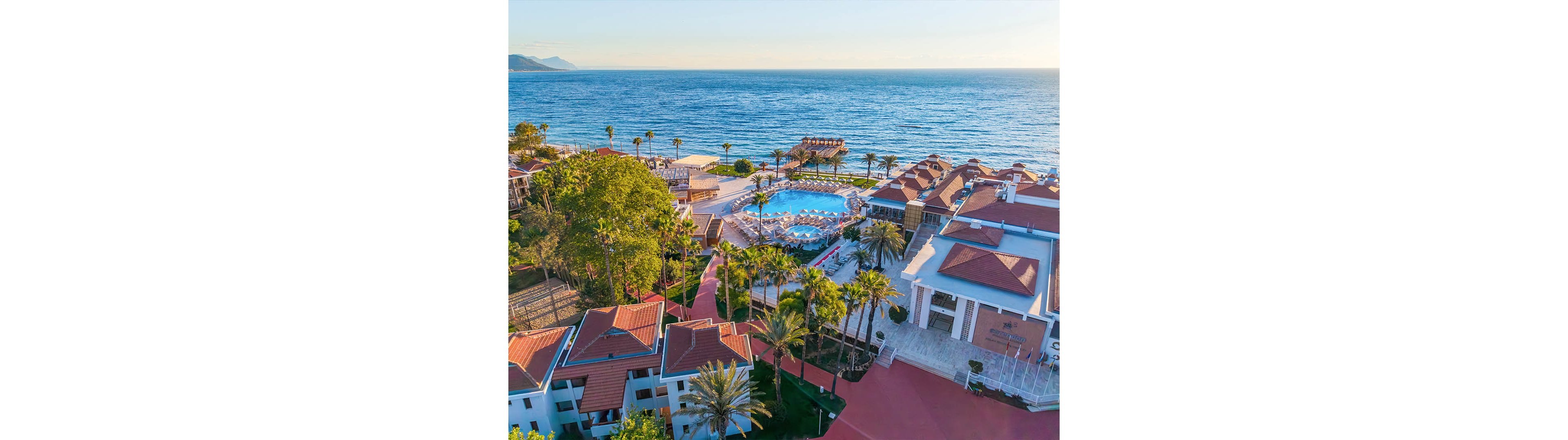 A seaside resort complex with red-roofed buildings is captured from above beside a calm blue ocean. At the center of the property, a large outdoor swimming pool is surrounded by palm trees and sun loungers. A wooden pier extends into the water, emphasizing the beachfront location. The scene is brightly lit with clear skies, suggesting a warm, relaxing vacation atmosphere.
