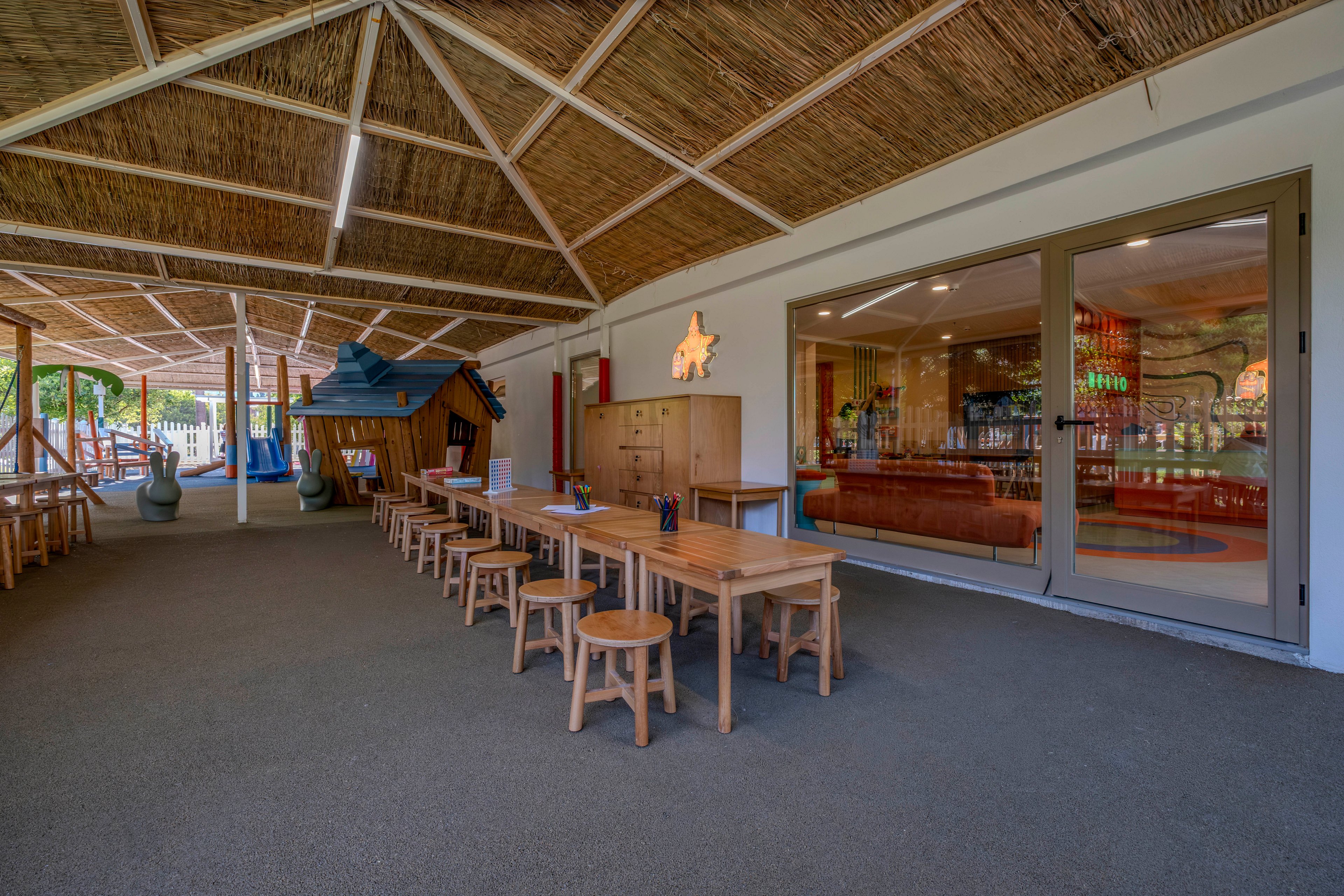 Spacious covered play area features long wooden tables and small stools arranged for children's activities. A whimsical wooden playhouse structure stands to the side, adding a playful focal point. Large glass doors reveal an indoor lounge or playroom with warm lighting and colorful decor. The setting feels bright, safe, and inviting for group crafts, learning, or supervised play.