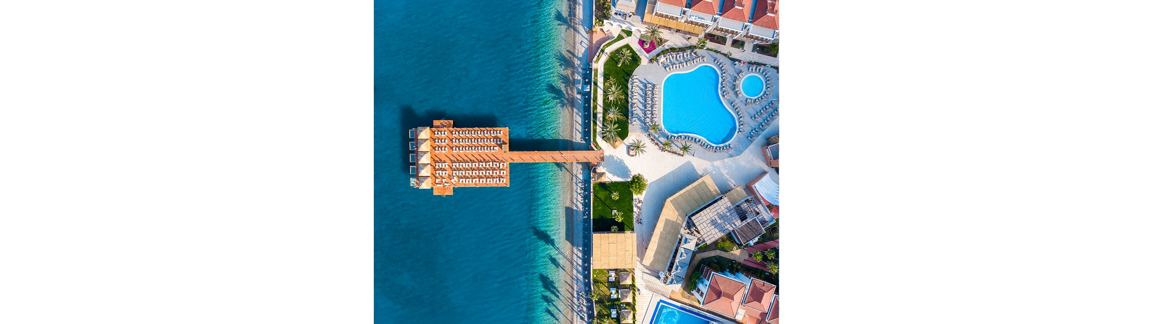 A coastal resort is captured from above, showing a long wooden pier extending into clear blue water. On shore, a large irregularly shaped swimming pool and a smaller circular pool are surrounded by sun loungers and palm trees. Nearby buildings with red-tiled roofs and additional pools create a dense vacation complex. The bright turquoise sea contrasts with the white pool decks and colorful structures, evoking a sunny holiday atmosphere.