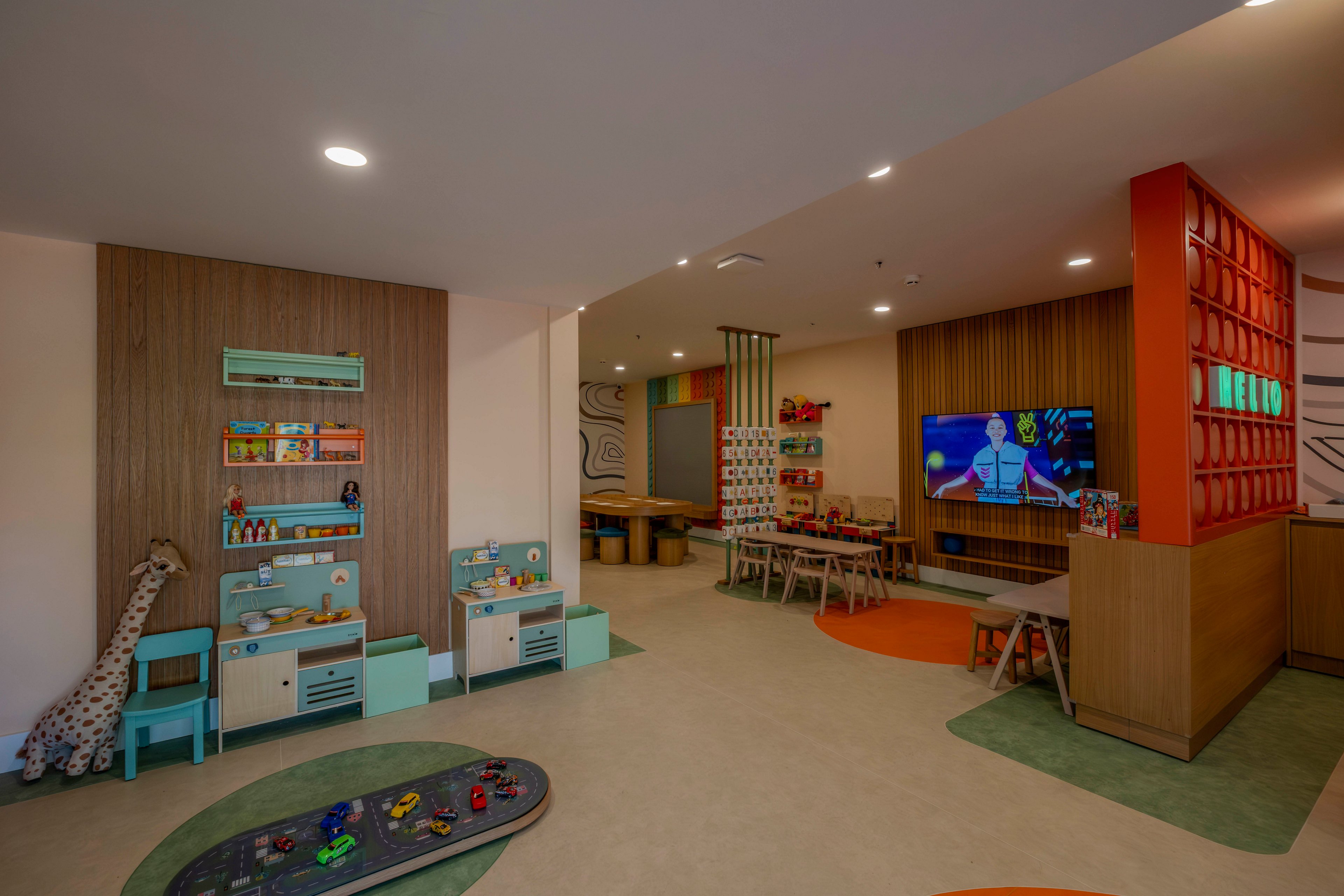 Spacious indoor children's playroom featuring low shelves filled with toys and books. A television screen displays an animated cartoon near a cluster of small tables and chairs. Soft play mats and colorful circular floor accents define different activity zones. Warm lighting and muted pastel colors create a cozy, inviting atmosphere for kids.