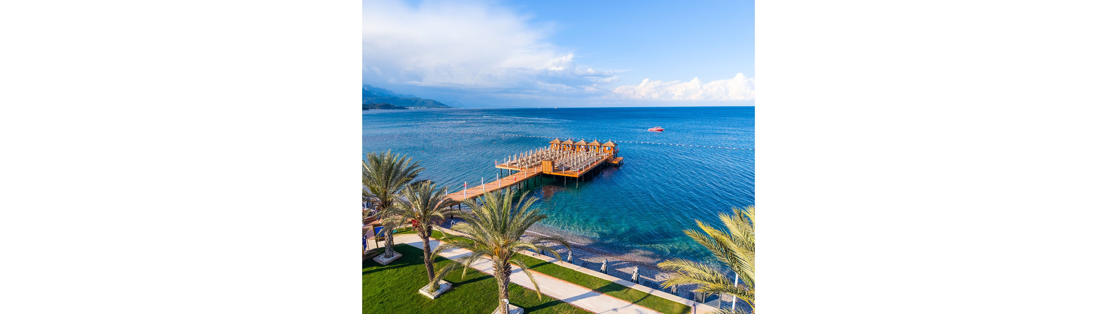 A wooden pier with rows of sun loungers and umbrellas extends over clear blue coastal water. Palm trees and manicured green lawns line the foreground along a stone walkway beside the beach. Gentle waves lap against the pebbled shoreline under a bright, partly cloudy sky. A small red boat floats in the distance, adding a pop of color to the tranquil seascape.