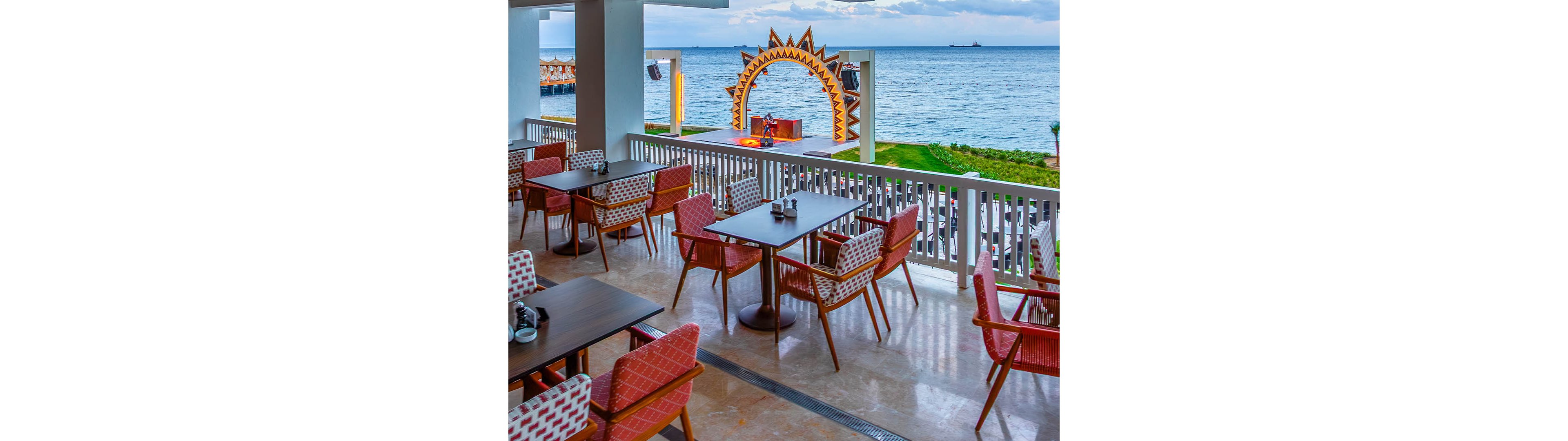 An open-air restaurant terrace overlooks a calm ocean with a decorative stage in the background. Empty tables and patterned chairs are neatly arranged along a white railing. The stage features a large sun-shaped arch and a performer near the shoreline. The scene suggests a relaxed coastal resort setting prepared for dining and entertainment.