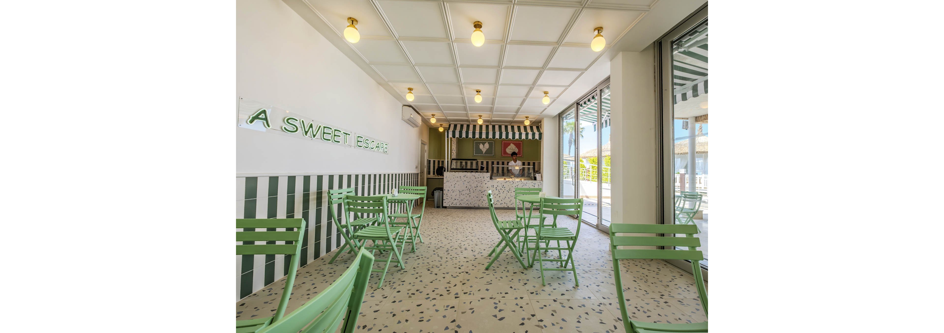 Modern dessert café interior with mint green metal chairs and small tables arranged across a terrazzo floor. The counter at the back features striped awnings and hanging pendant lights, creating a playful, retro-inspired look. Large windows on the right let in natural light and reveal an outdoor area. A neon-style wall sign reads "A SWEET ESCAPE," reinforcing the cozy, inviting atmosphere.