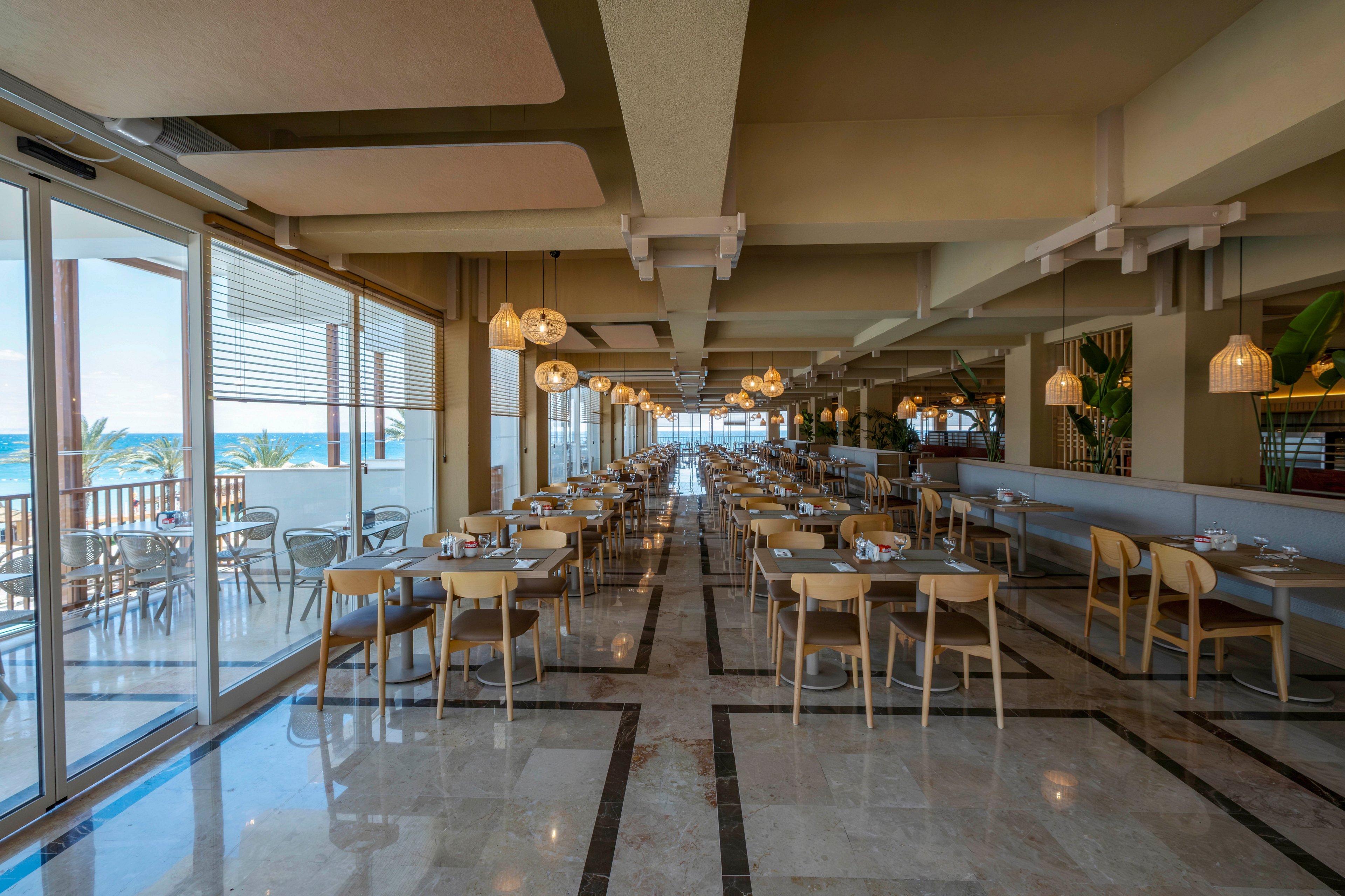 Spacious restaurant dining room with rows of wooden tables and chairs is shown in warm natural light. Large windows and glass doors on the left open to a balcony with views of the sea. Pendant lights hang from the ceiling, reflecting softly on the polished marble floor. The setting appears calm and ready for guests, with neatly arranged place settings on each table.