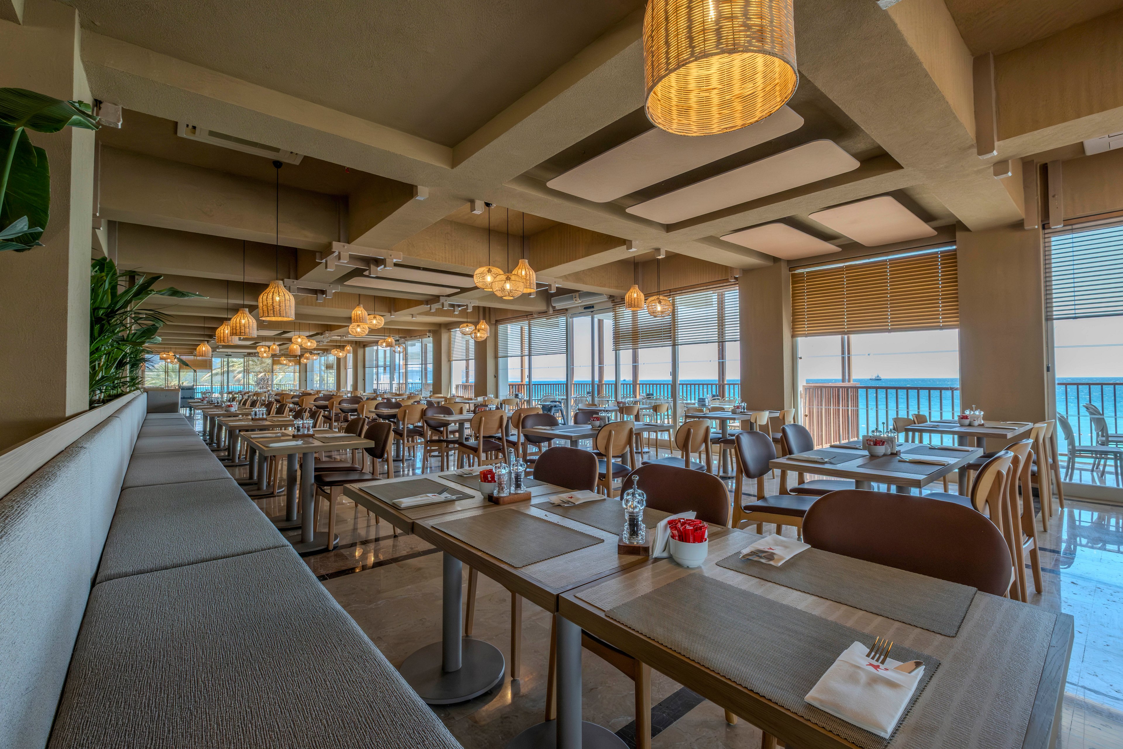 Spacious contemporary restaurant interior featuring rows of neatly set wooden tables and chairs. Large windows line the side of the room, offering a clear view of the sea and bright natural light. Warm pendant lights and neutral-toned decor create a cozy yet modern atmosphere. The tables are prepared with placemats, napkins, cutlery, and small condiment holders, ready for guests.