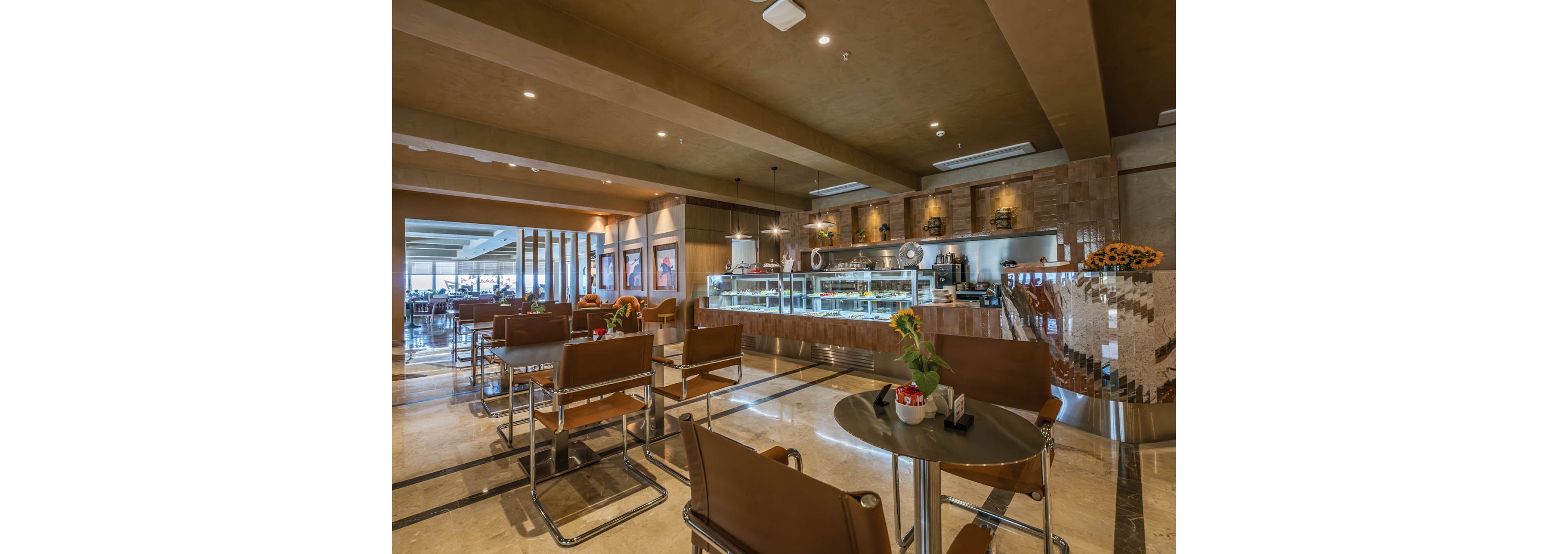 A contemporary café or restaurant interior features neatly arranged tables and brown leather chairs. The central focus is a well-lit service counter with glass display cases and coffee equipment. Warm lighting, tiled walls, and polished floors create an inviting, upscale atmosphere. Sunflowers and small table decorations add a touch of color to the neutral palette.