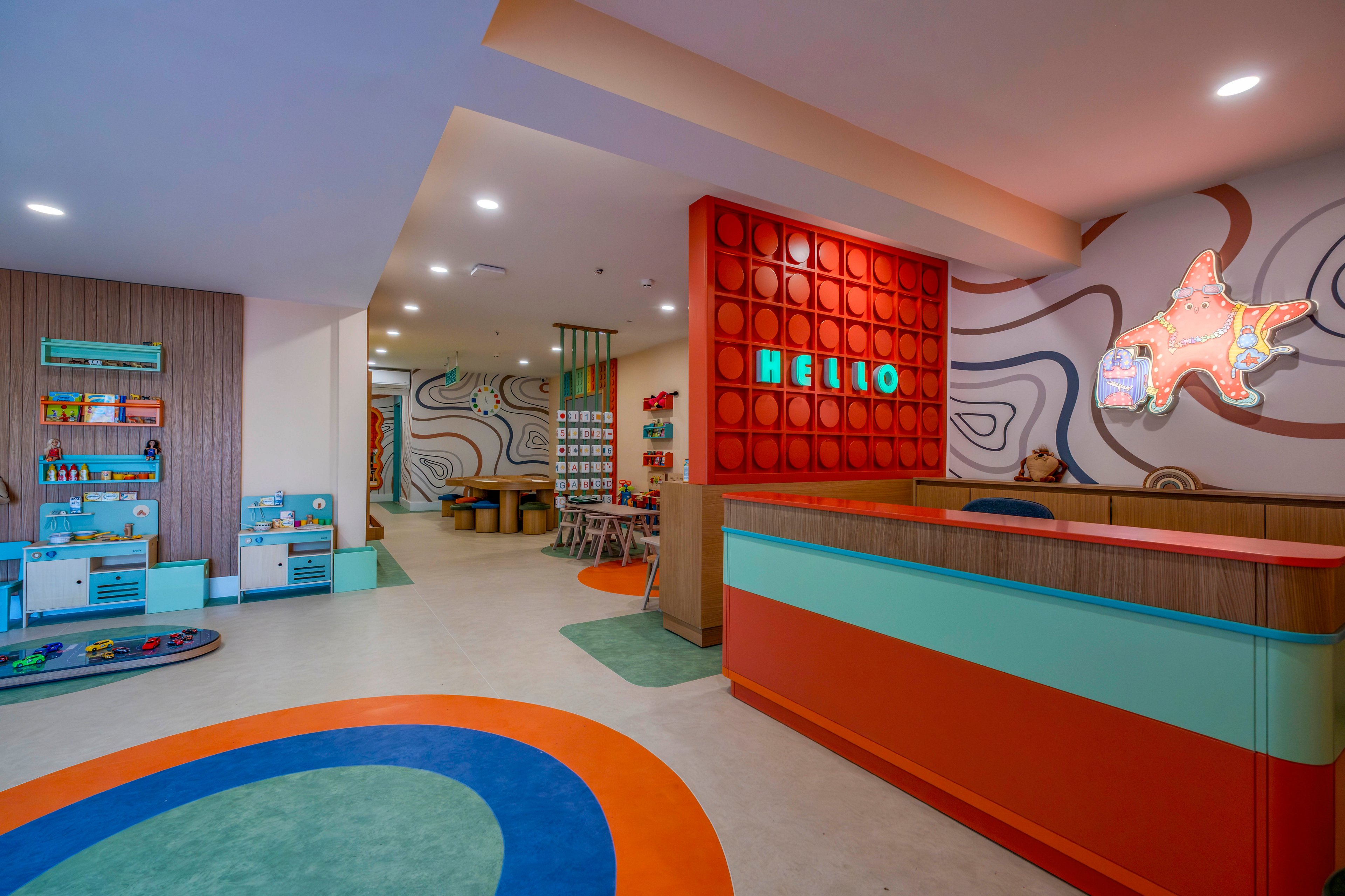 Brightly colored children's playroom features a modern reception desk in the foreground. The wall behind the desk displays the word HELLO in illuminated letters on a red geometric panel. In the background, play tables, shelves with toys, and wall-mounted activity boards create an engaging environment. Curved patterns on the walls and floor add a playful, contemporary design. Soft lighting and open space make the room inviting and child-friendly.