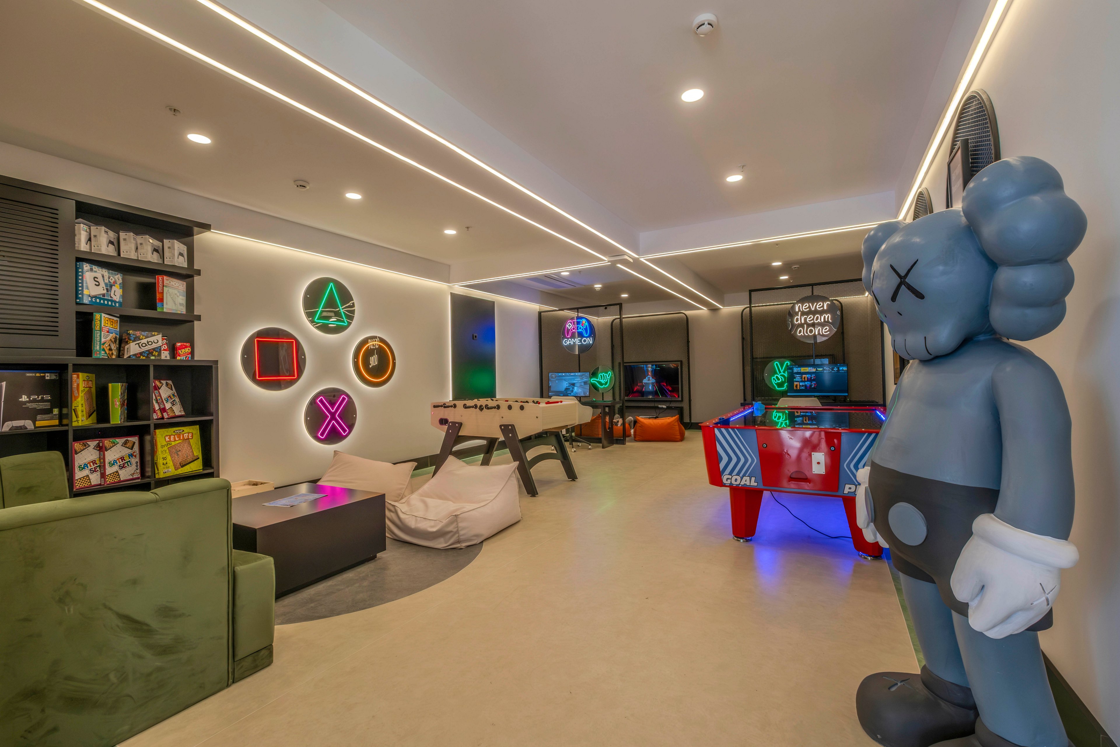A contemporary gaming lounge features a mix of arcade machines, a foosball table, and a colorful air hockey table. Neon wall art with geometric controller symbols adds a playful glow to the room. Comfortable seating, beanbags, and shelves filled with books and collectibles create a relaxed hangout atmosphere. A large stylized character statue stands prominently near the entrance, enhancing the creative, youthful vibe.