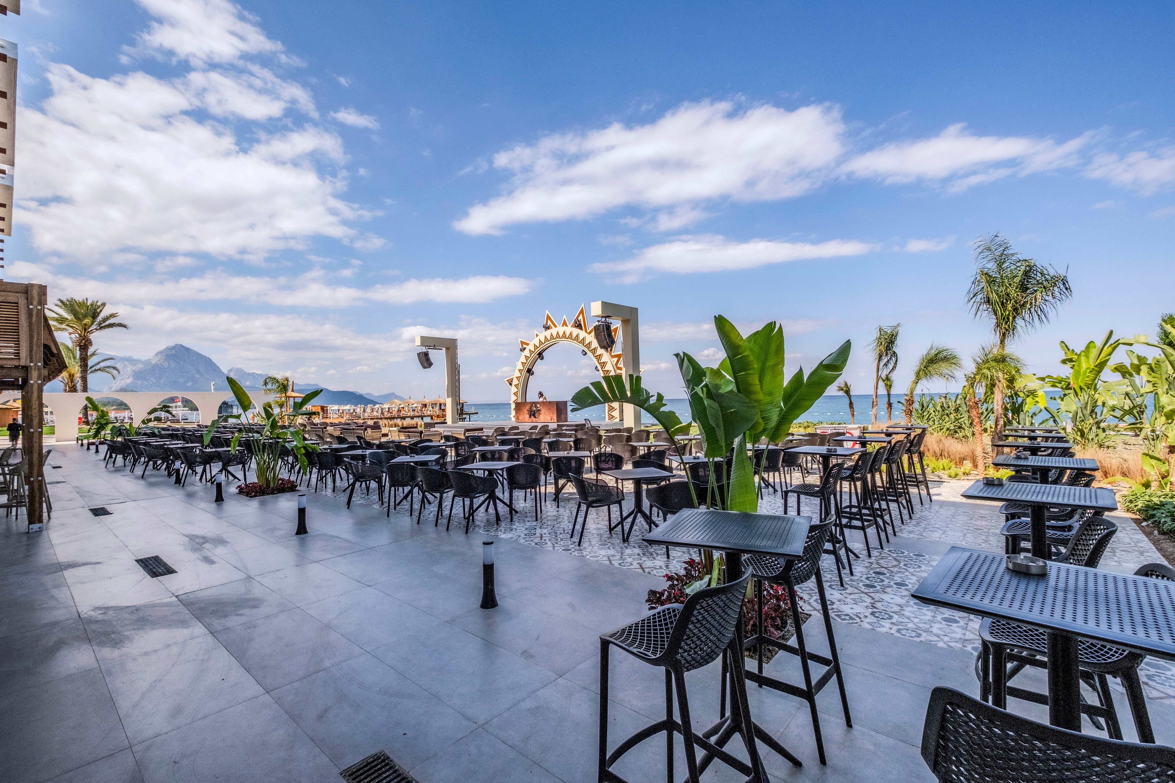 An expansive outdoor café terrace is arranged with rows of black tables and chairs. The setting overlooks the sea, framed by a decorative archway and distant mountains under a bright blue sky. Tropical plants and palm trees line the edges of the patio, adding lush greenery to the scene. The atmosphere feels relaxed and inviting, ideal for seaside dining or refreshments.