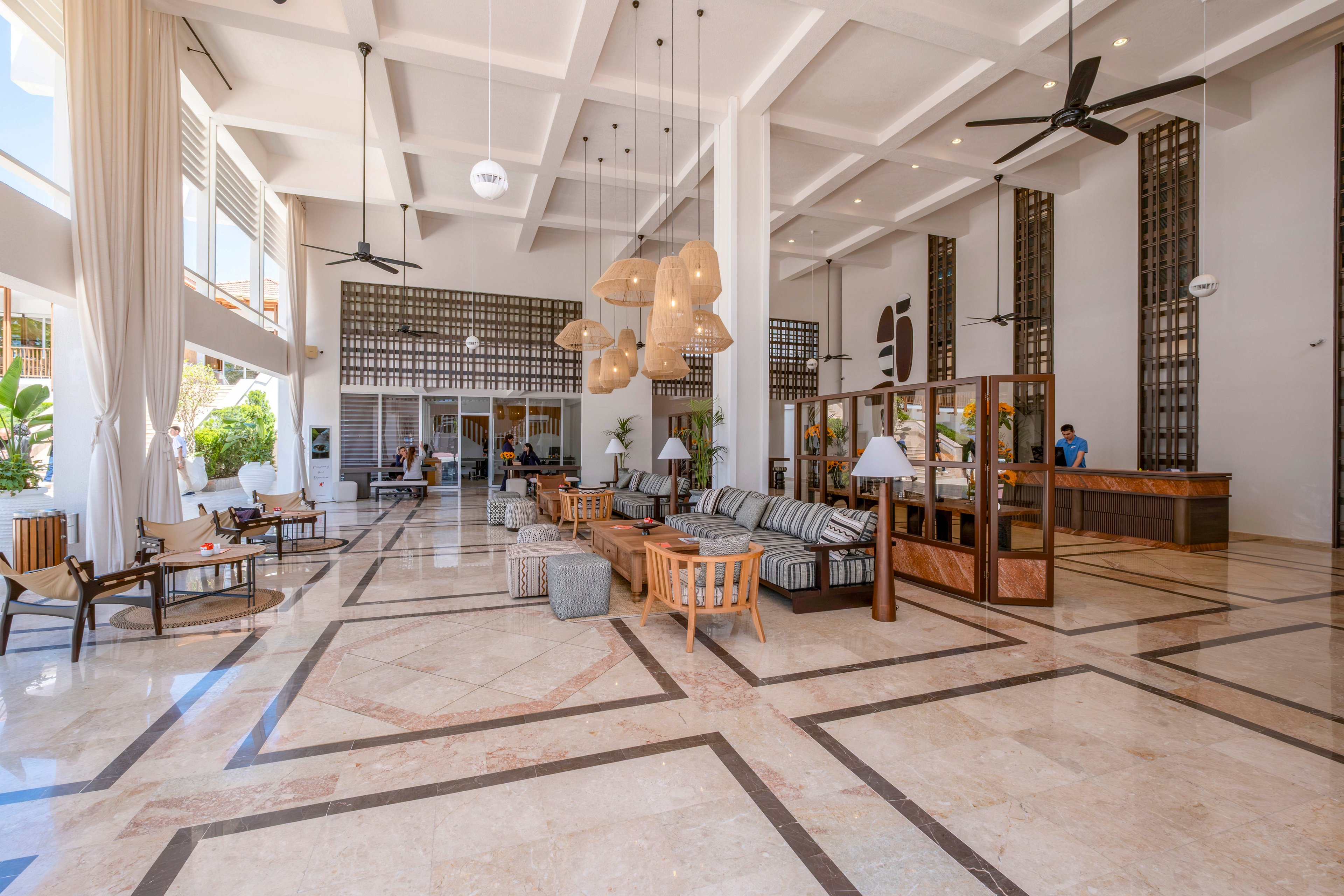 A large, airy hotel lobby features polished marble floors and high ceilings with exposed beams. Contemporary lounge seating and wooden furniture are arranged in cozy conversation areas. Pendant lights and ceiling fans hang above, while floor-to-ceiling windows and sheer curtains let in natural light. A reception desk is visible on the right, with staff assisting guests in the bright, welcoming space.