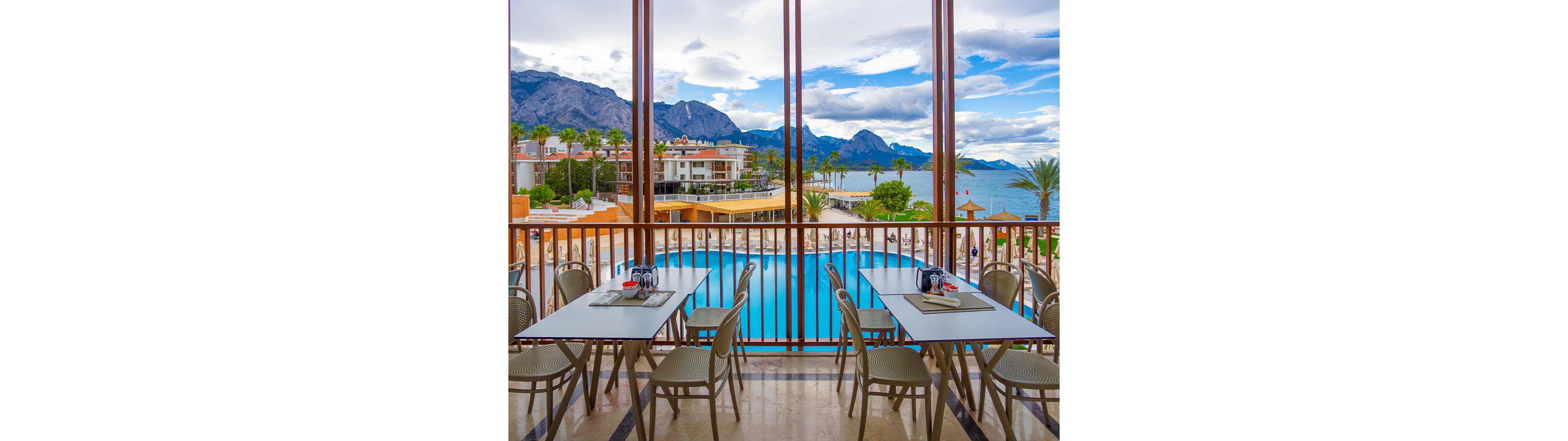 An outdoor resort terrace features neatly arranged dining tables and chairs overlooking a large blue swimming pool. Beyond the pool, palm trees and low-rise hotel buildings sit along a scenic coastline. Majestic mountains rise in the background under a partly cloudy sky, creating a dramatic natural backdrop. The scene suggests a relaxed vacation atmosphere with a focus on leisure, dining, and waterfront views.