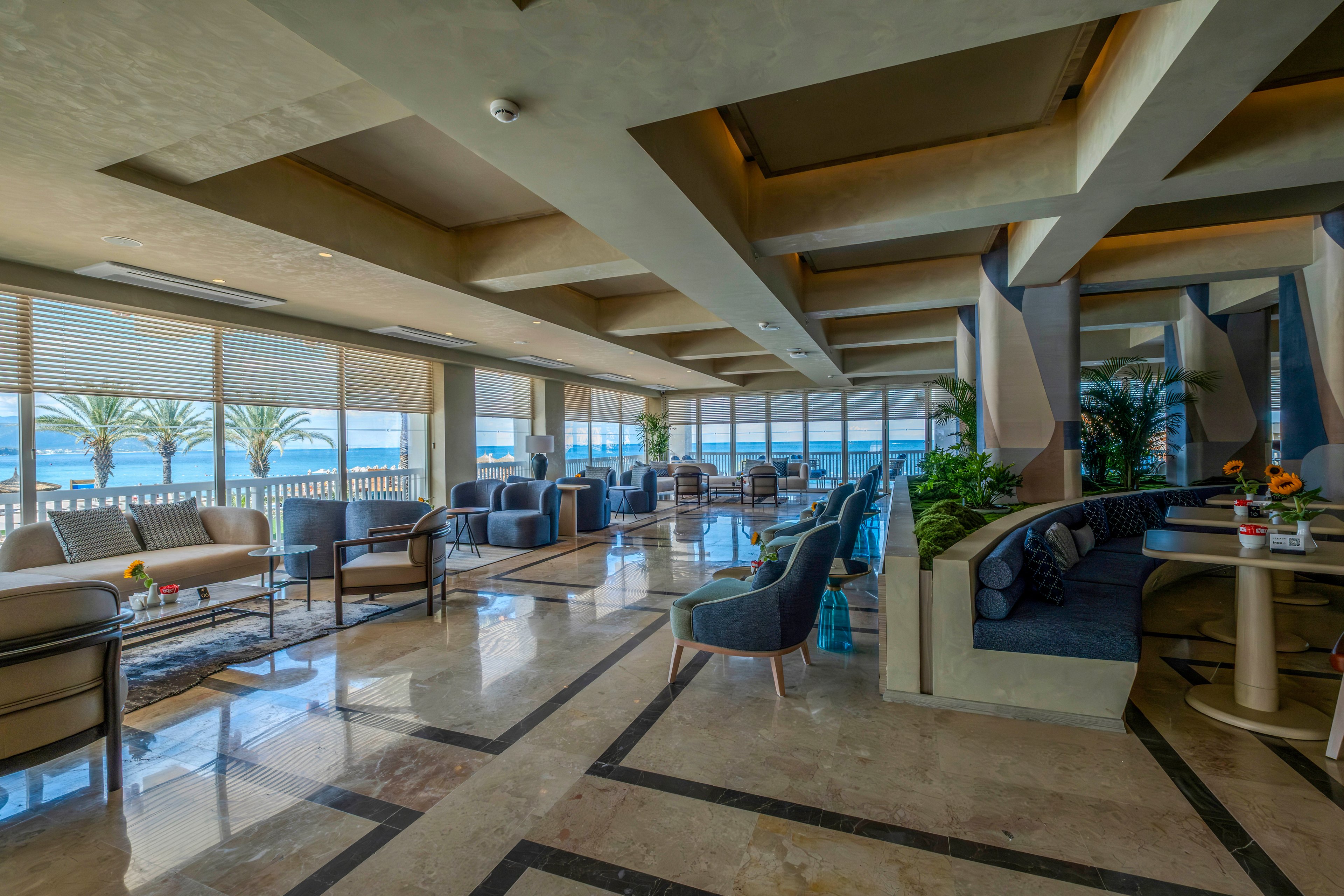 Spacious contemporary lounge area features comfortable sofas and armchairs arranged in conversational groupings. Large floor-to-ceiling windows reveal a bright seaside view with palm trees and calm water outside. Polished marble floors and geometric ceiling details enhance the upscale, modern design. Potted plants, side tables, and small floral arrangements add warmth and color to the relaxing coastal setting.
