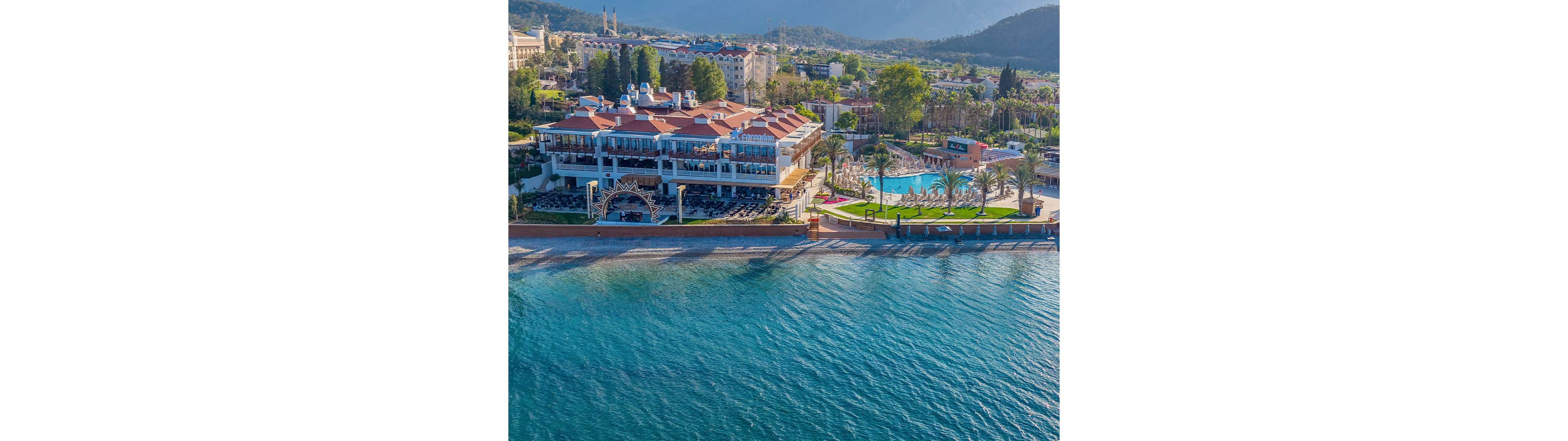 A large seaside resort hotel sits directly on a clear blue shoreline. The complex features multiple red-roofed buildings, an outdoor swimming pool, and palm-lined walkways. Sun loungers and umbrellas are arranged along the waterfront and around the pool area. In the background, additional buildings and green hills create a scenic coastal landscape.
