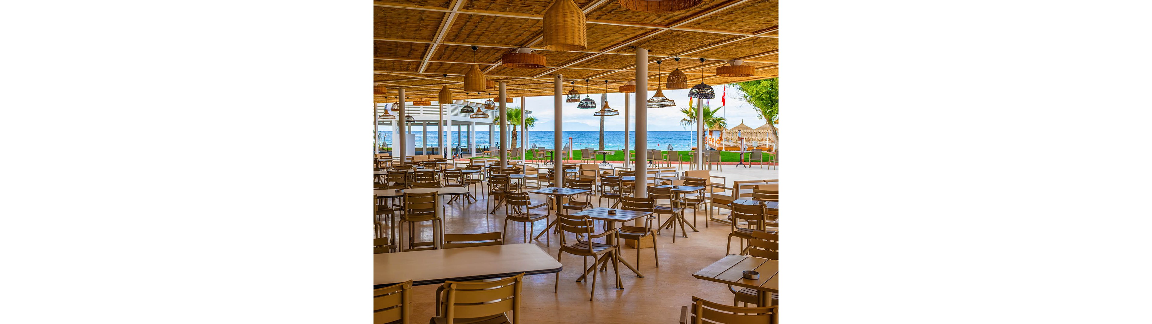 An open-air seaside restaurant features rows of empty tables and chairs under a woven, natural-style ceiling. Wicker pendant lights hang above the seating area, creating a relaxed coastal atmosphere. Beyond the terrace, palm trees and green lawn lead to a sandy beach and calm blue sea. The scene suggests a tranquil vacation setting before guests arrive.