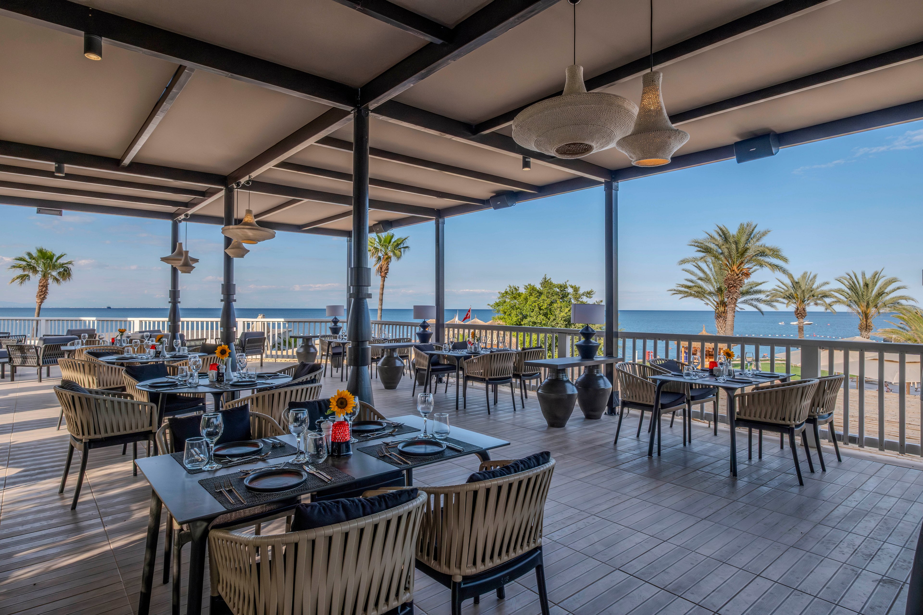An elegant open-air restaurant terrace overlooks a calm blue sea. Neatly arranged tables with woven chairs, glassware, and simple place settings create a refined dining atmosphere. Decorative pendant lights hang from the ceiling, and large vases line the railing. Palm trees and clear skies in the background emphasize the coastal, vacation-like setting.