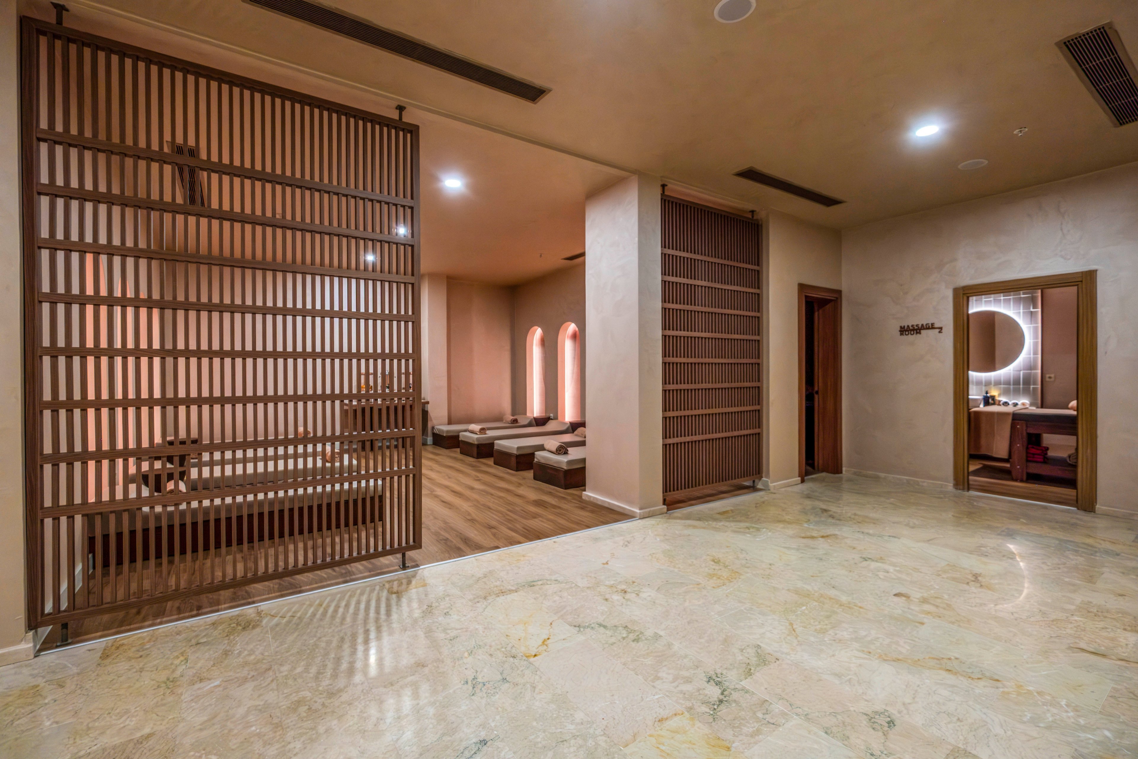 A contemporary spa interior features a spacious relaxation area with low cushioned loungers arranged along a wooden floor. Vertical wooden slat partitions divide the open space, adding privacy and architectural interest. Warm recessed lighting and soft beige walls create a calm, soothing atmosphere. A vanity or treatment room with a round illuminated mirror is visible through an open doorway in the background.