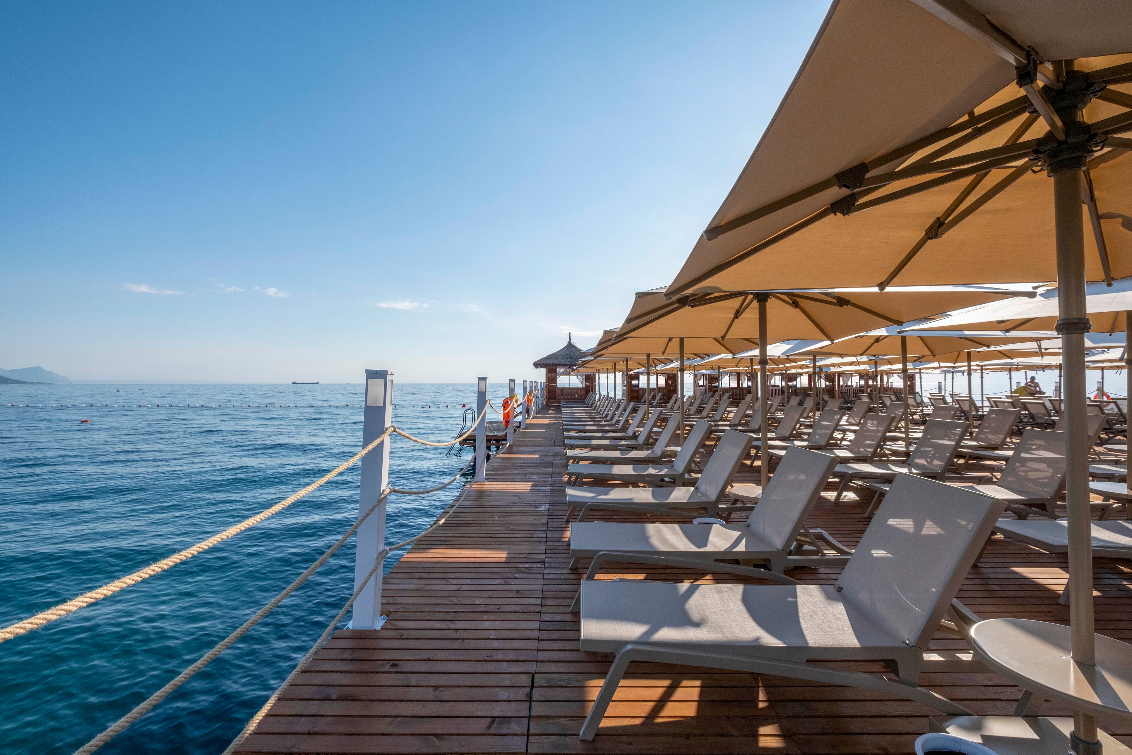 A row of empty sun loungers lines a wooden pier extending over calm blue water. Large beige umbrellas provide shade along the deck, creating a relaxed seaside atmosphere. The clear sky and gentle waves suggest a warm, sunny day at a coastal resort. The scene feels peaceful and inviting, with no people present on the pier.