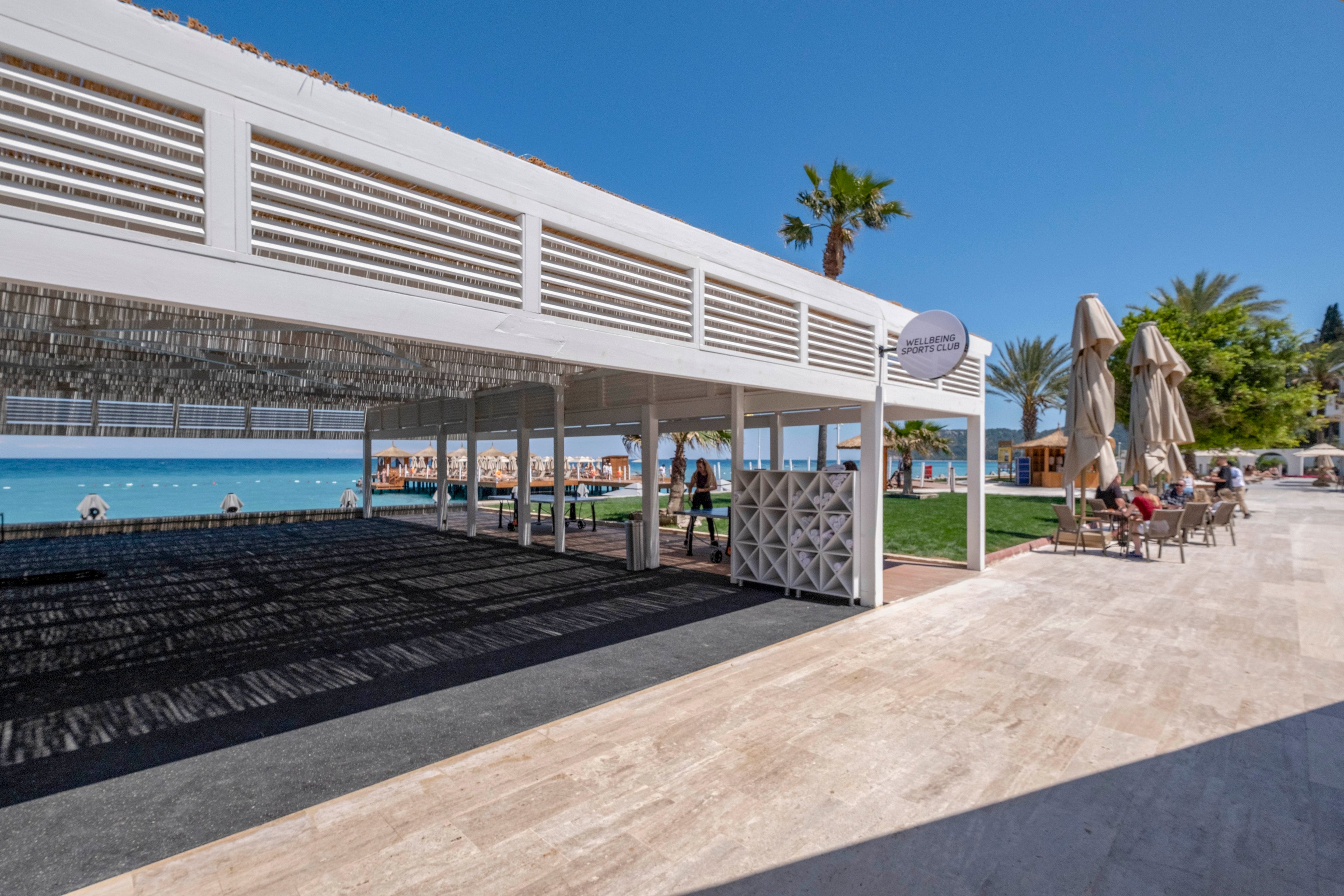 A modern white pavilion stands beside a seaside resort terrace on a bright sunny day. The scene overlooks a calm blue sea with sun loungers and umbrellas along the beach. Guests sit at outdoor café tables under large beige umbrellas near palm trees and manicured grass. The overall atmosphere is relaxed and summery, with clear skies and strong natural light.