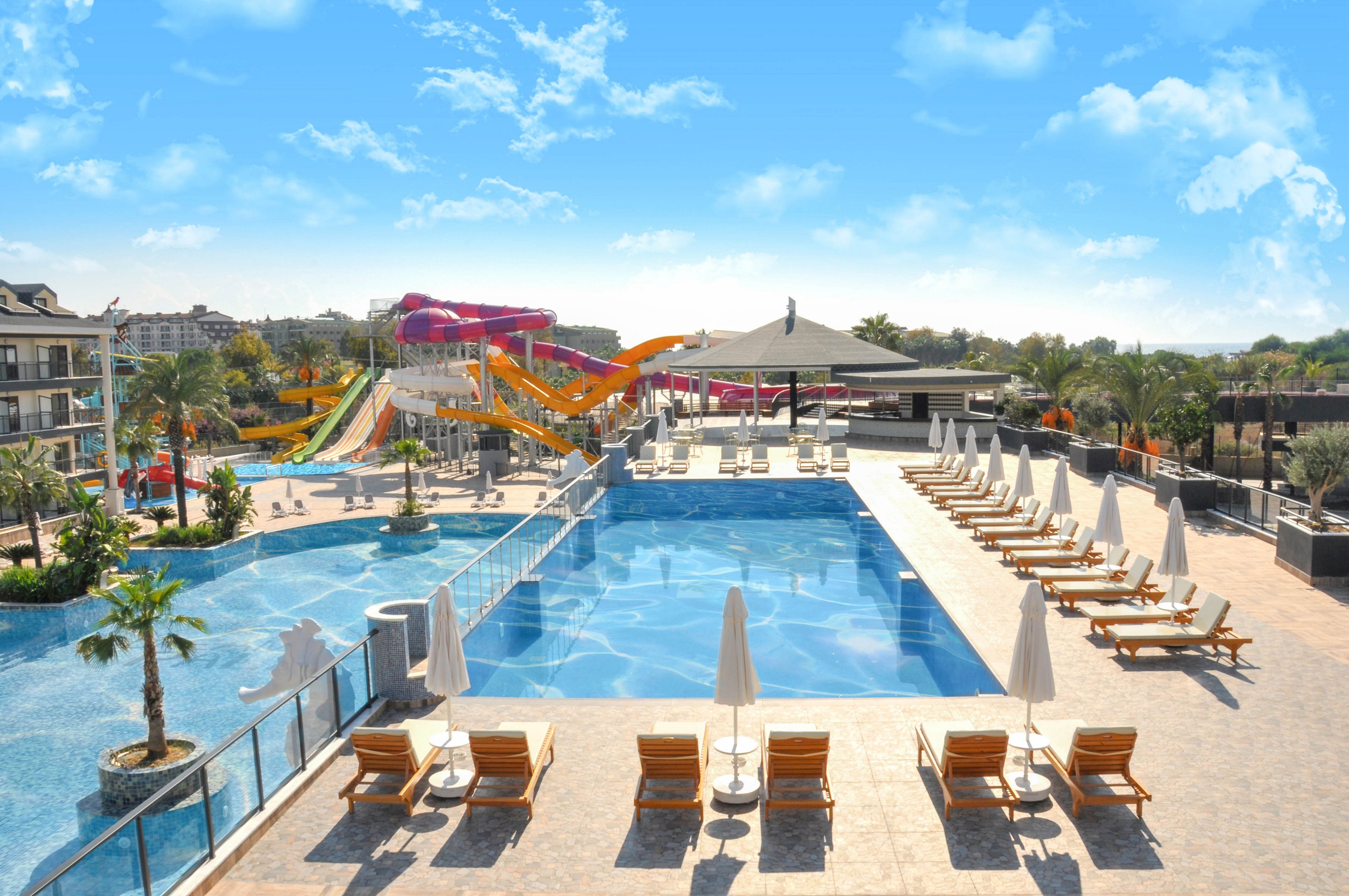 Large outdoor swimming pools are shown at a sunny resort with clear blue skies overhead. Numerous lounge chairs with white umbrellas line the pool deck, suggesting a relaxing vacation setting. In the background, several tall, colorful water slides curve into an adjacent pool area, adding a playful, family-friendly atmosphere. Palm trees and modern buildings surround the pools, enhancing the tropical, leisure-focused environment.