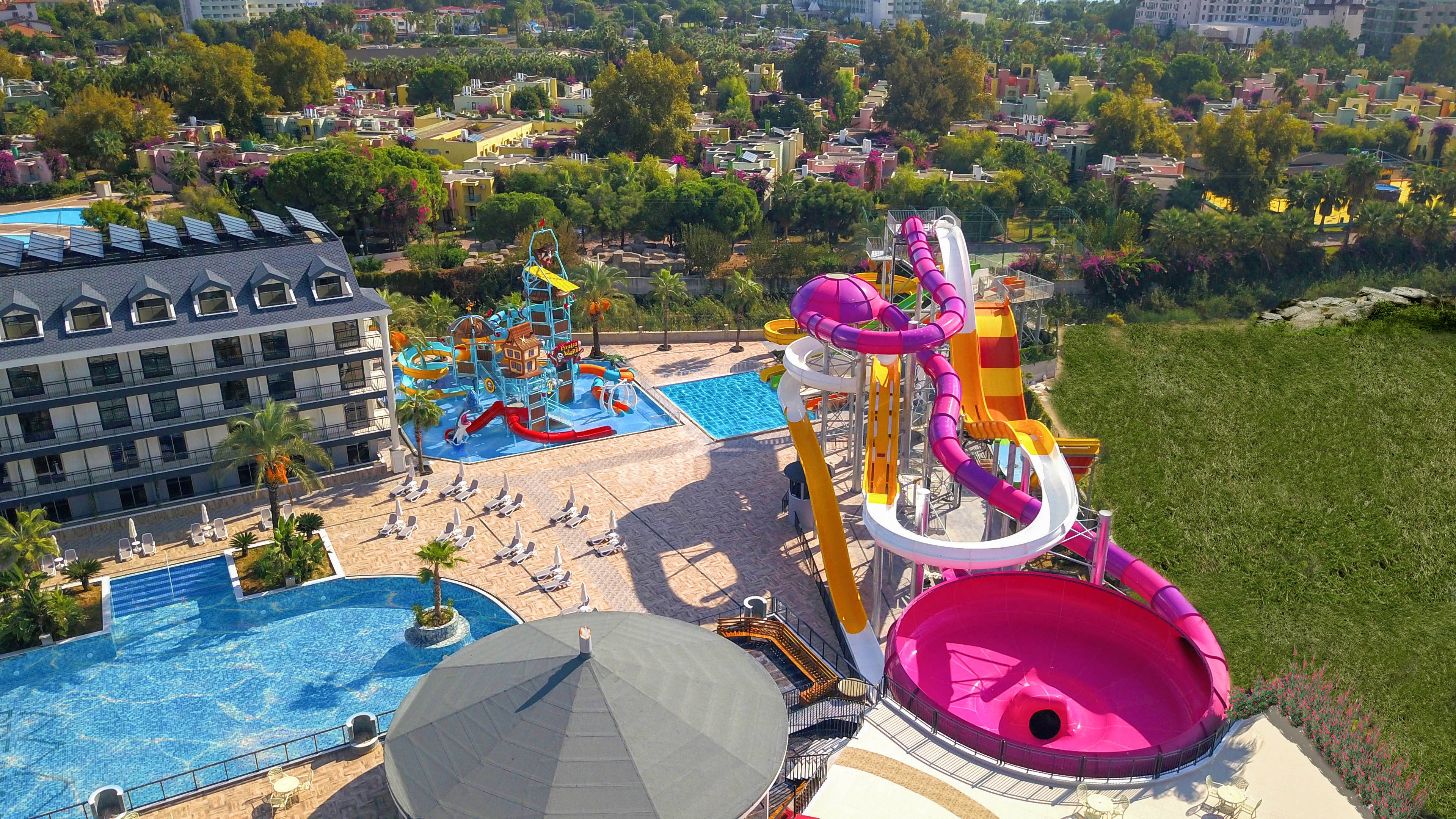 A large resort waterpark is captured from above, showcasing multiple pools and bright waterslides. A towering spiral slide with pink and white tubes ends in a big circular splash bowl. Nearby, a children’s play area features smaller slides, fountains, and climbing structures surrounded by sun loungers. A modern hotel building borders the pool deck, while lush greenery and additional buildings stretch into the background.