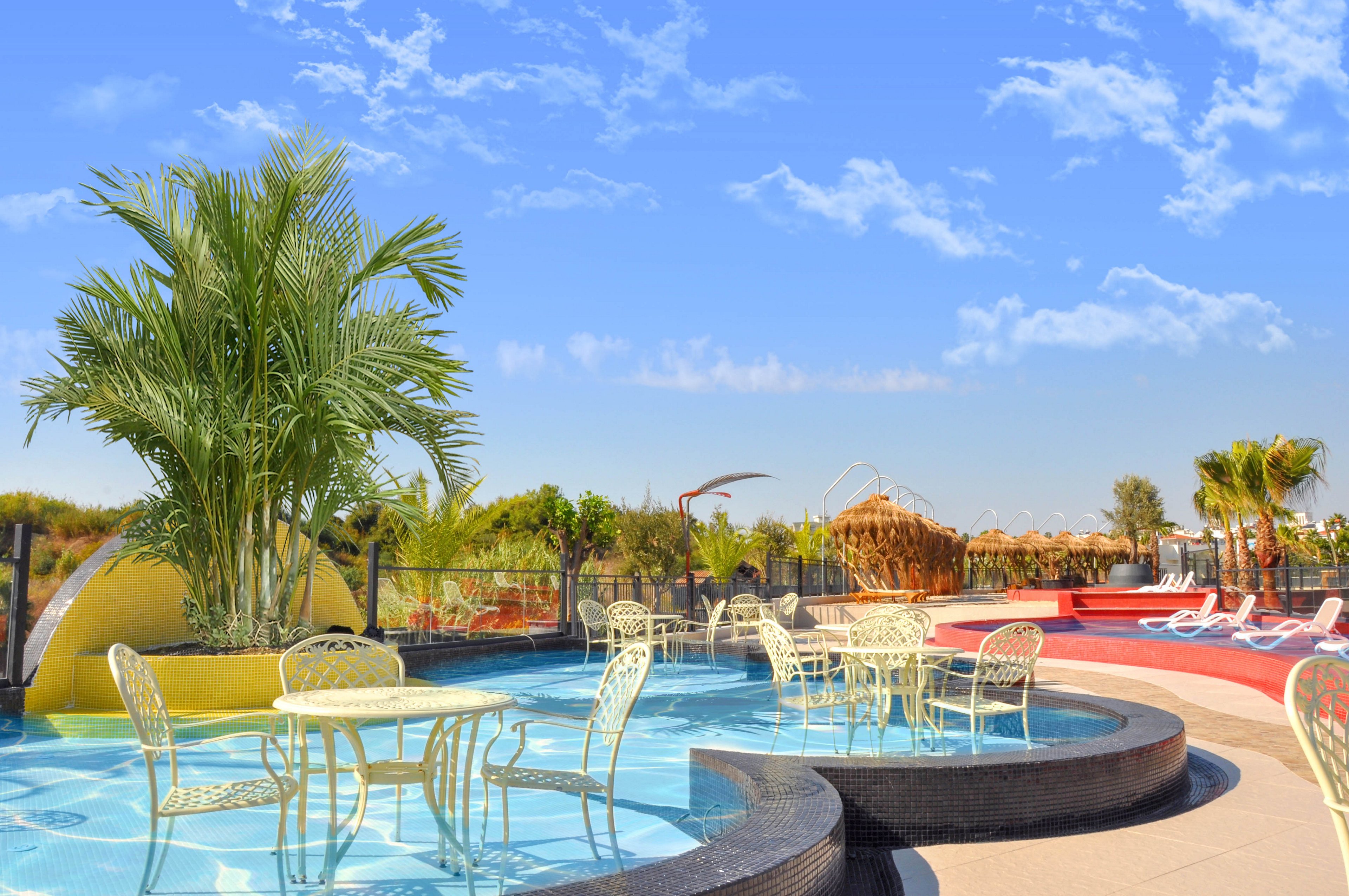 An outdoor resort pool features curved, tiered basins with clear blue water and decorative palm plants. Light-colored metal tables and chairs are placed in the shallow water, creating a unique seating area. In the background, sun loungers line the poolside under a bright blue sky with scattered clouds. The scene suggests a warm, relaxing vacation atmosphere in a landscaped leisure area.
