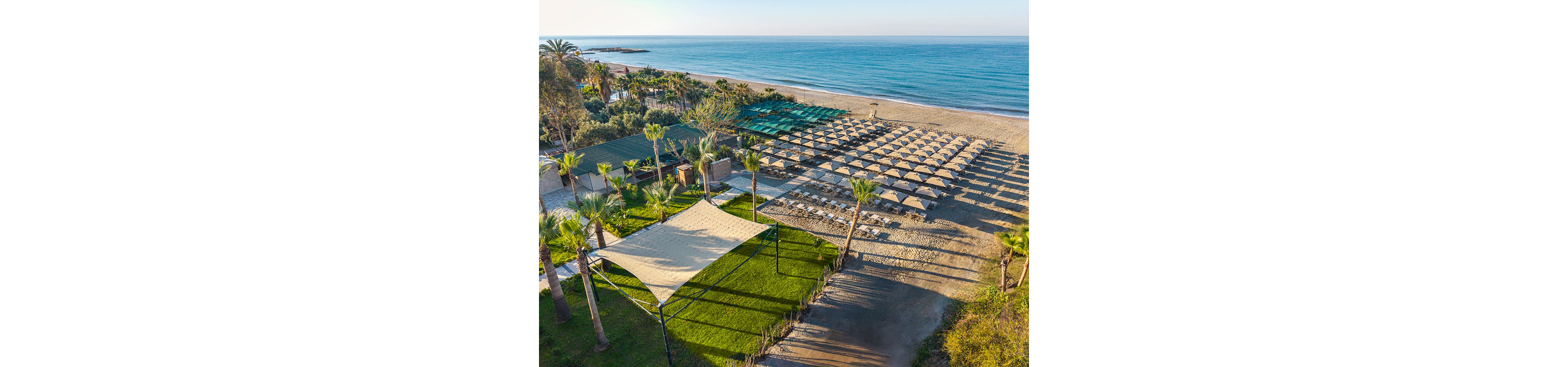 Aerial view of a sunny seaside resort with rows of sun loungers and umbrellas neatly arranged on a sandy beach. The calm blue sea stretches to the horizon under a clear sky. Shaded canopy areas and green lawns with palm trees provide comfortable relaxation spaces near the shoreline. The scene suggests a peaceful vacation destination with well-maintained facilities and ample seating.