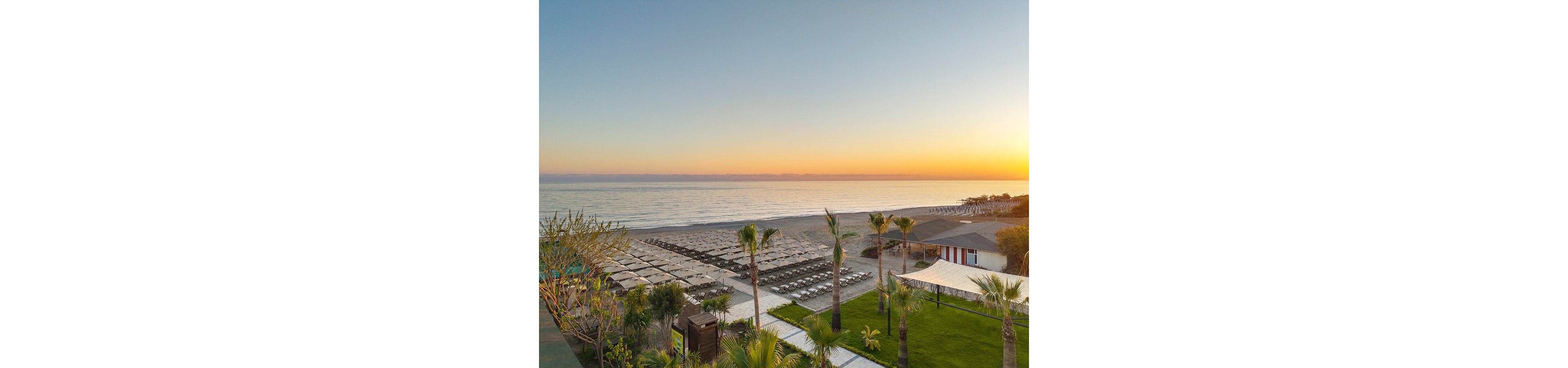 A serene beachfront resort is captured at sunrise with the sun low on the horizon over calm sea waters. Neatly arranged rows of sun loungers and umbrellas line the sandy beach, stretching toward the shoreline. In the foreground, manicured green lawns, palm trees, and landscaped plants frame the view. A small building and shaded pavilion sit near the beach, enhancing the organized, relaxing holiday atmosphere.