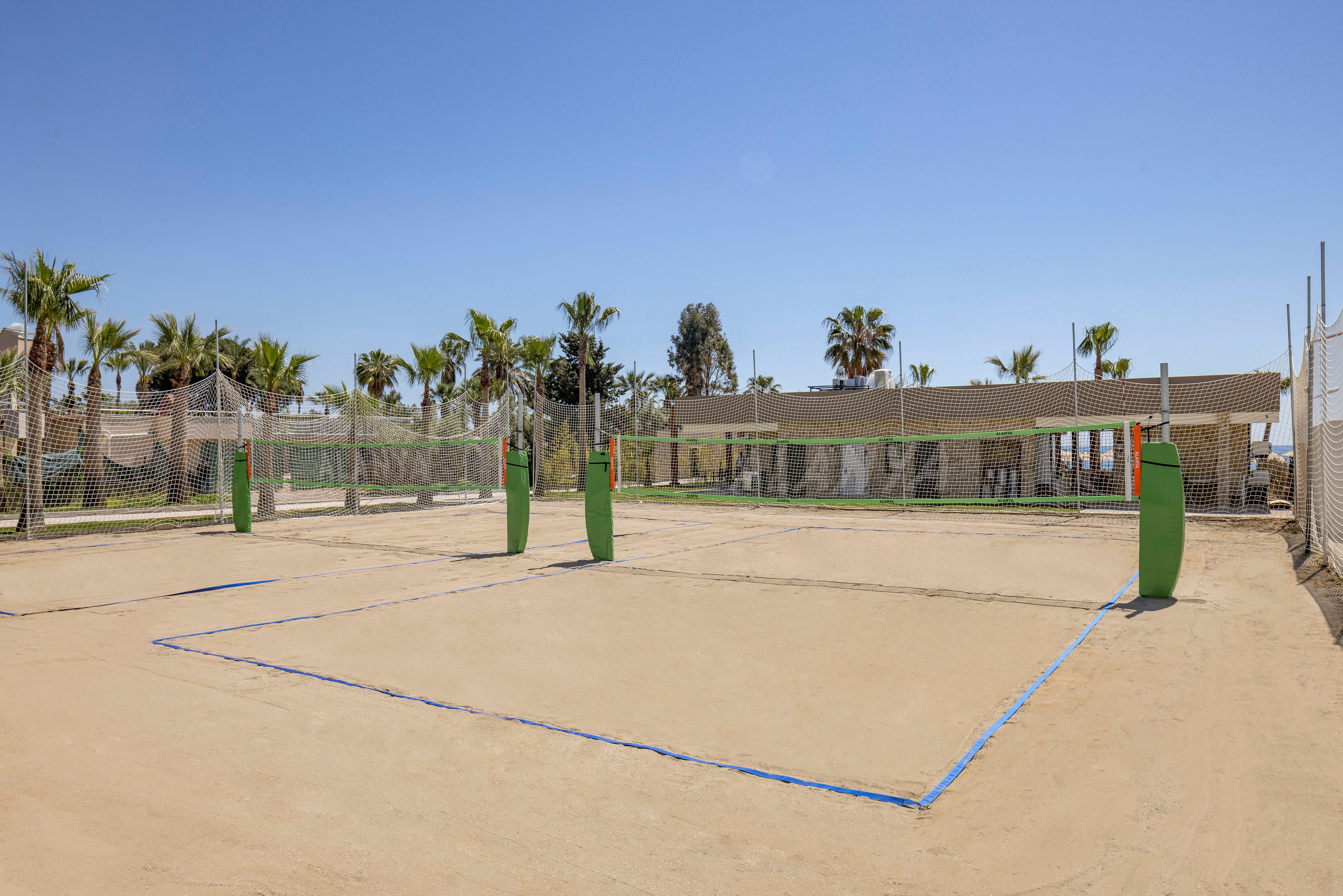A pair of empty beach volleyball courts sit on smooth sand under a bright blue sky. Nets are stretched between green-padded posts, with blue boundary lines marking the playing areas. Palm trees and a low building are visible in the background, suggesting a resort or recreational facility. The scene feels sunny, open, and ready for outdoor sports activity.