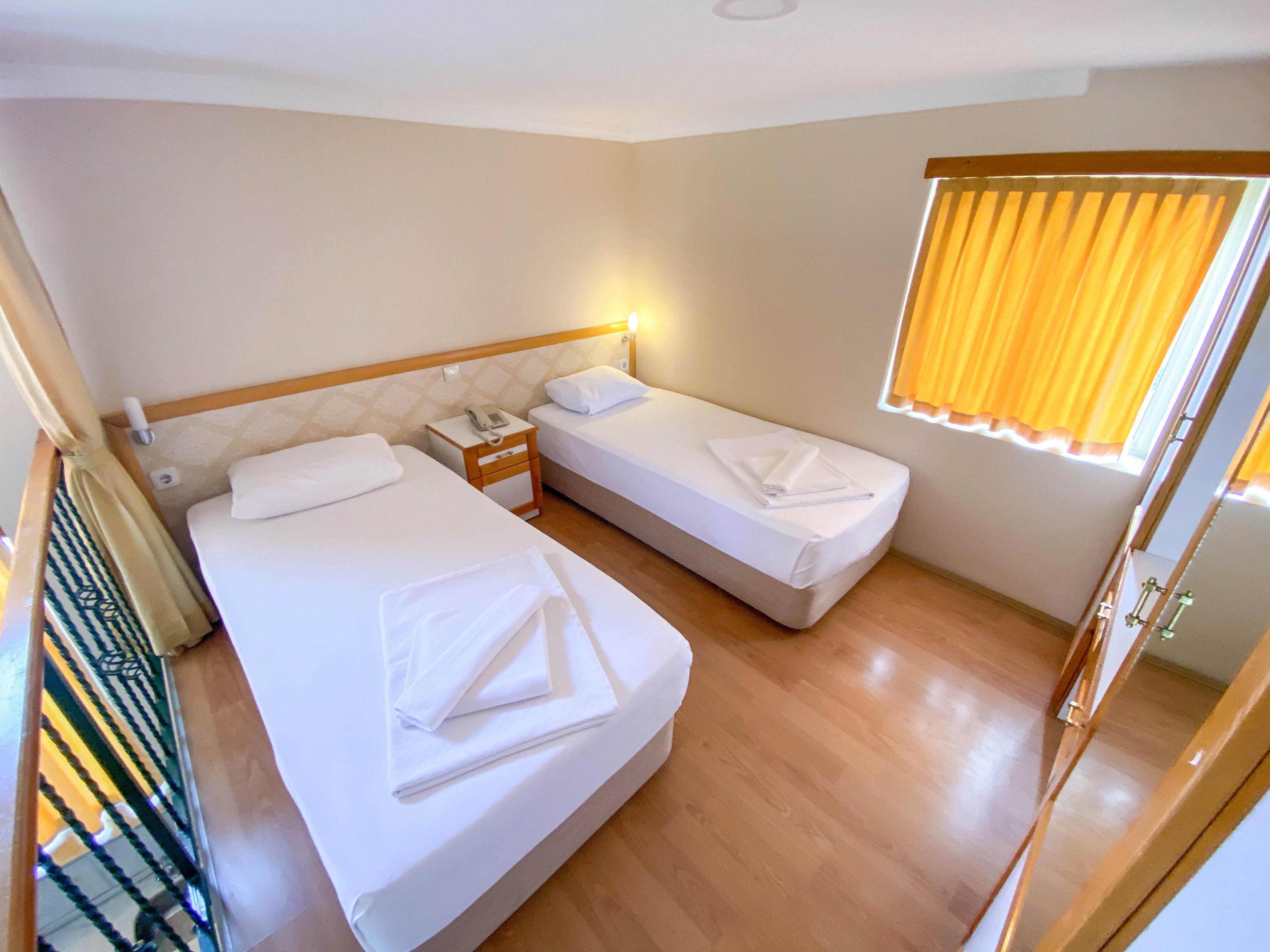 A compact hotel room features two neatly made single beds with white linens and folded towels. The space has light-colored walls and a warm wooden floor, creating a clean and simple atmosphere. Sunlight filters through yellow curtains covering a window, adding a soft glow to the room. A small bedside table with a lamp sits between the beds, emphasizing a functional and minimalist design.