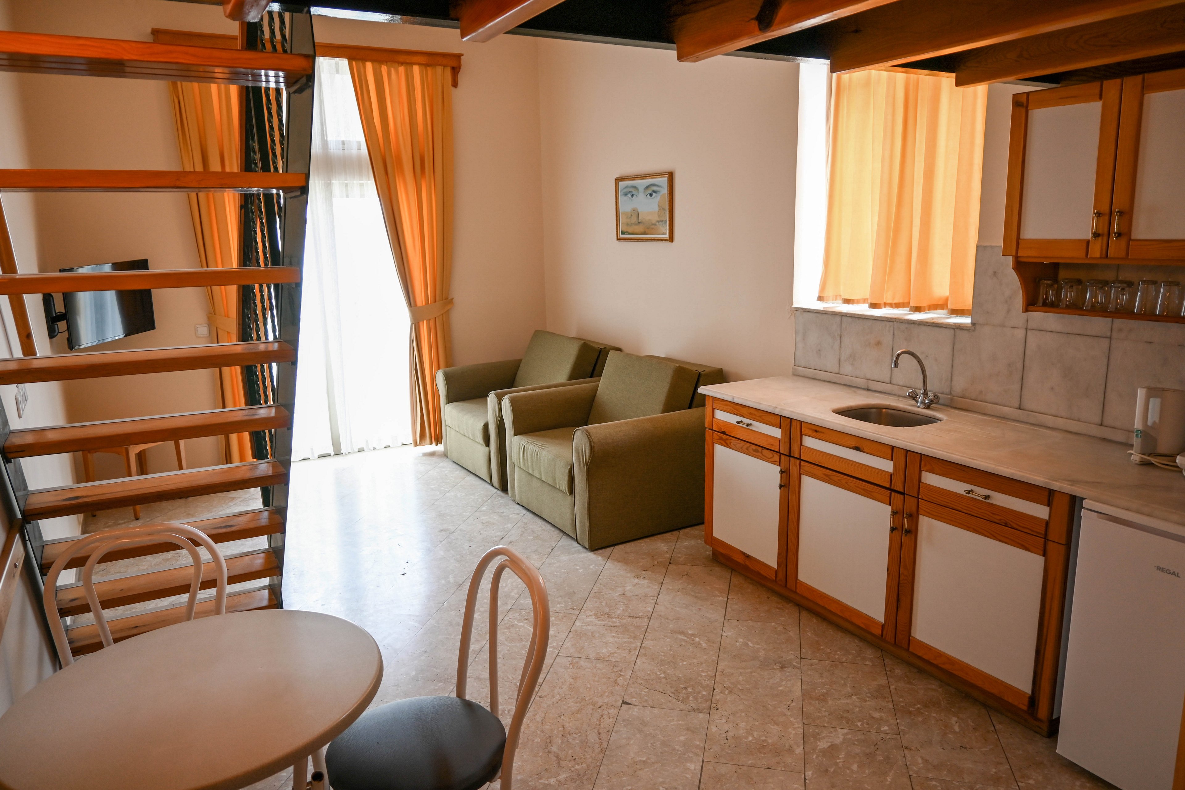 Compact loft-style apartment interior featuring a small kitchenette and seating area. A green sofa sits against the wall near tall windows with sheer curtains and orange drapes. Wooden cabinets, a sink, and tiled backsplash define the kitchen space, while a round table with chairs provides a dining spot. An open wooden staircase leads to an upper level, adding vertical interest to the bright, sunlit room.