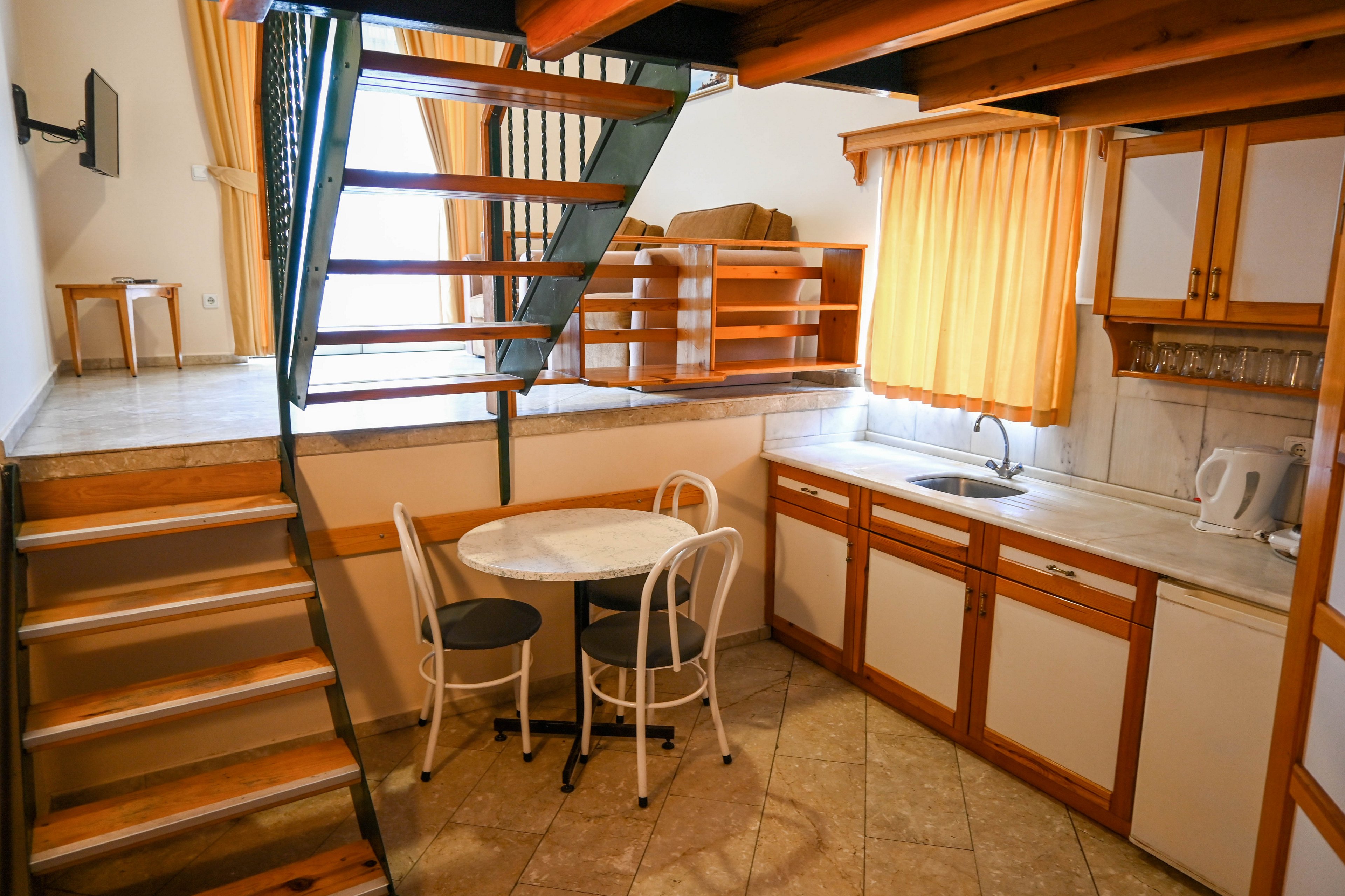 Compact loft-style apartment interior featuring a small kitchen and dining area. A metal and wood staircase leads up to an elevated sleeping or living platform. The kitchen includes wooden cabinets, a sink, kettle, and glassware, with a round white table and three chairs in the center. Warm yellow curtains and tiled flooring create a bright, inviting atmosphere.