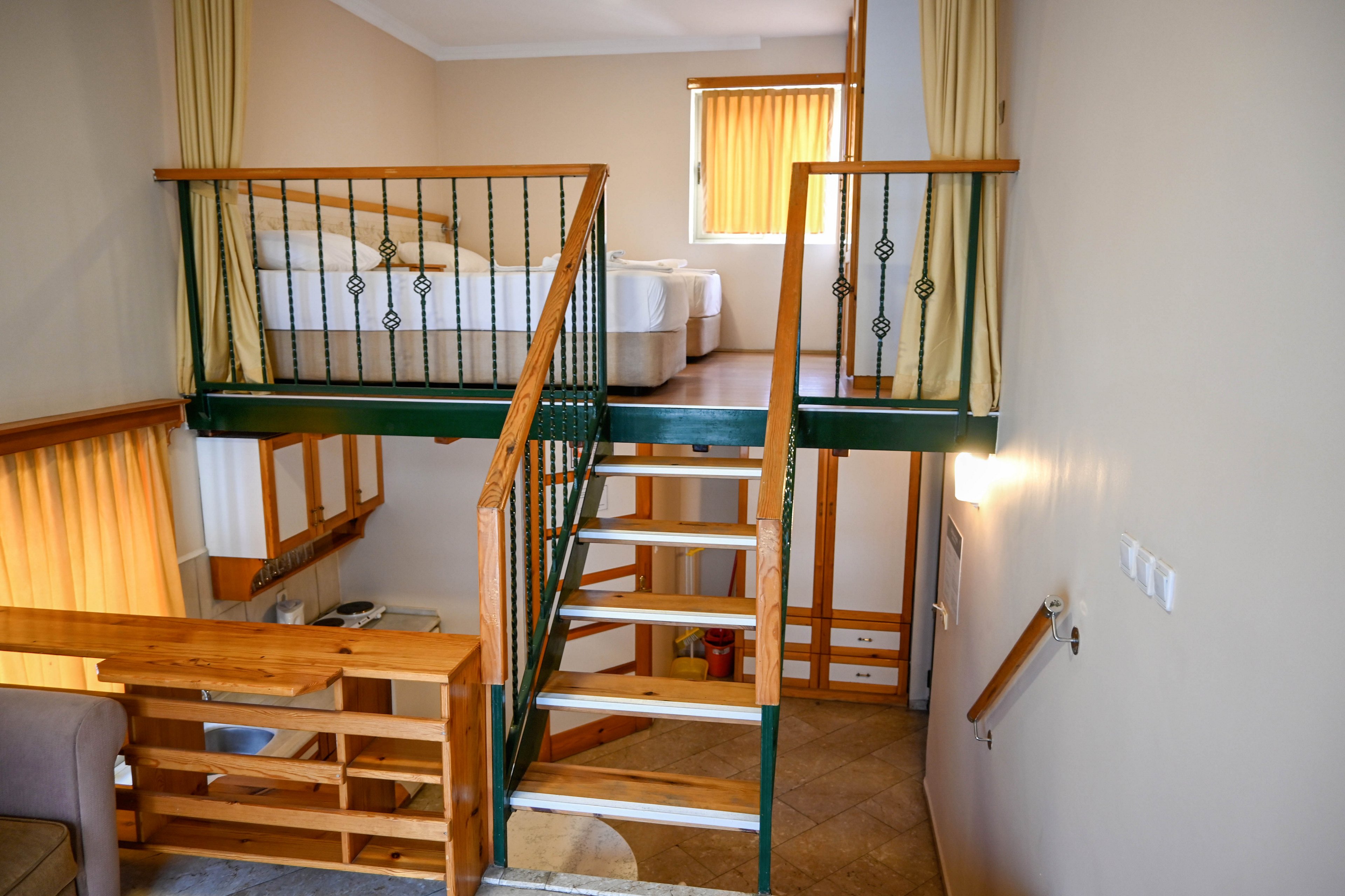 Interior view of a modern loft-style hotel room featuring a mezzanine sleeping area with two single beds. A central staircase with metal railings connects the upper level to the lower living space. Warm beige walls, wooden accents, and soft yellow curtains create a cozy atmosphere. Natural light filters through vertical blinds on the windows, highlighting the clean and minimalist design.
