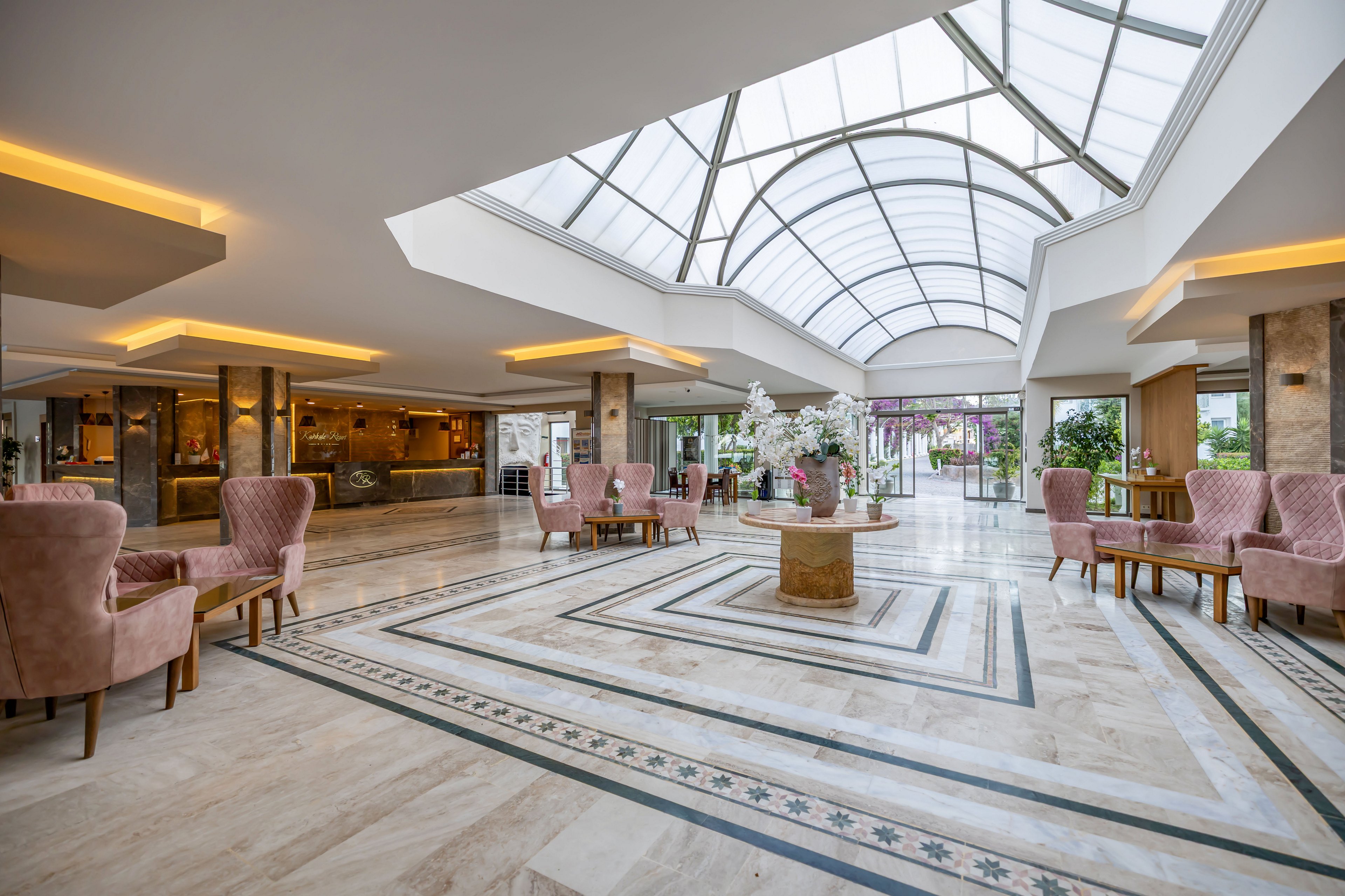 Bright, expansive hotel lobby featuring a large arched glass ceiling and polished marble flooring. Pink upholstered armchairs and small tables are arranged in seating areas along both sides of the space. A round central table displays a floral arrangement, adding elegance to the modern interior. Warm recessed lighting and floor-to-ceiling windows at the far end create an inviting atmosphere with views of greenery outside.