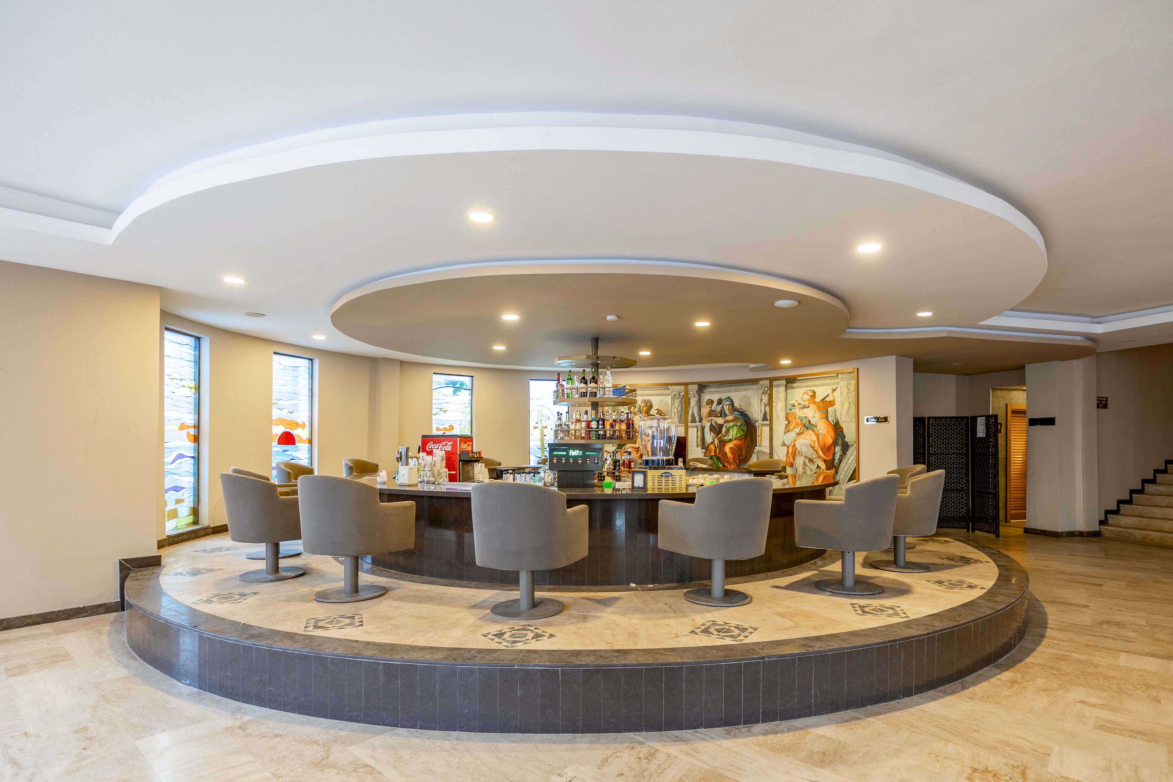 A contemporary circular bar counter sits at the center of a spacious hotel lobby. Several swivel armchairs surround the bar, facing shelves of bottles and glassware. Large decorative murals and tall windows add color and visual interest to the neutral-toned space. Recessed ceiling lights and polished flooring create a clean, inviting atmosphere suitable for guests and social gatherings.