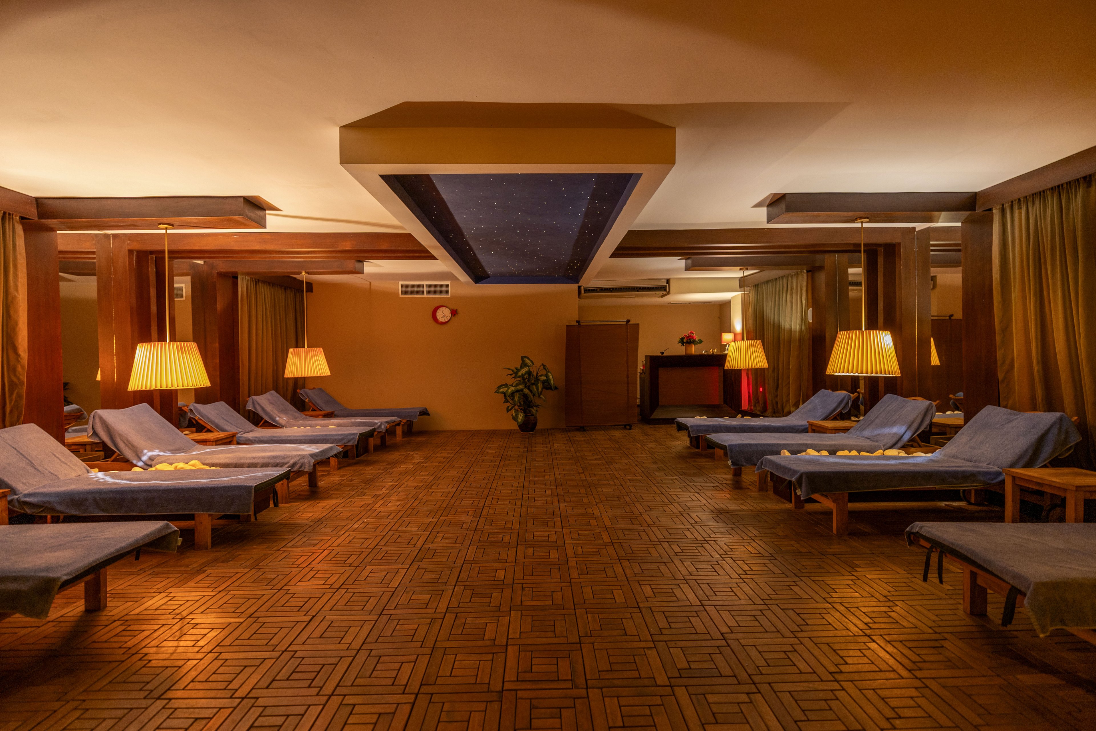 A tranquil spa relaxation lounge features rows of cushioned loungers covered with blue towels. Soft, warm lighting from table lamps creates a cozy atmosphere across the wooden parquet floor. The room is empty, emphasizing calm and privacy, with a plant and reception area visible in the background. Neutral brown and gold tones dominate the space, enhanced by the ceiling detail and draped curtains.