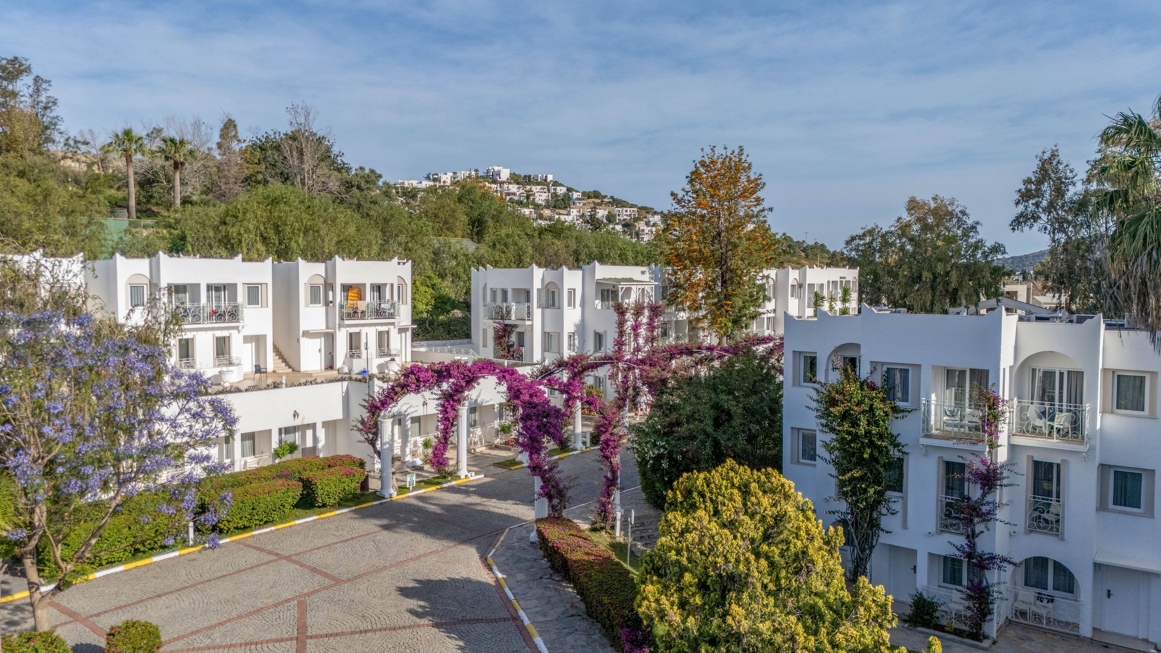 A cluster of white, low-rise apartment buildings is surrounded by lush greenery and flowering trees. The scene features a paved courtyard and driveway leading through the complex, with purple bougainvillea arching over the path. Hillside residences are visible in the background under a clear blue sky, enhancing the sense of a peaceful residential neighborhood. The overall atmosphere is bright, sunny, and inviting.