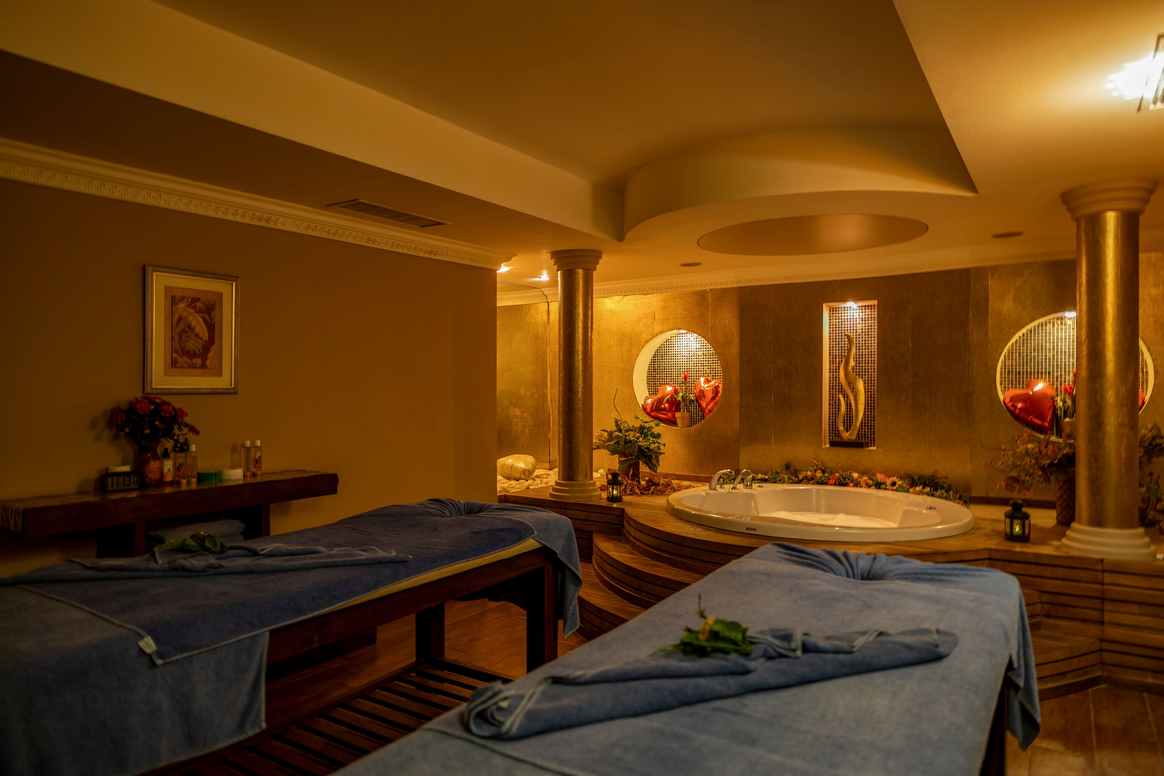 A warmly lit spa room features two massage tables covered with blue sheets, ready for treatments. In the background, a large whirlpool tub is framed by golden columns and decorative wall art. Soft ambient lighting and earth-toned walls create a relaxing, intimate atmosphere. Fresh flowers and neatly arranged towels add to the sense of comfort and wellness.