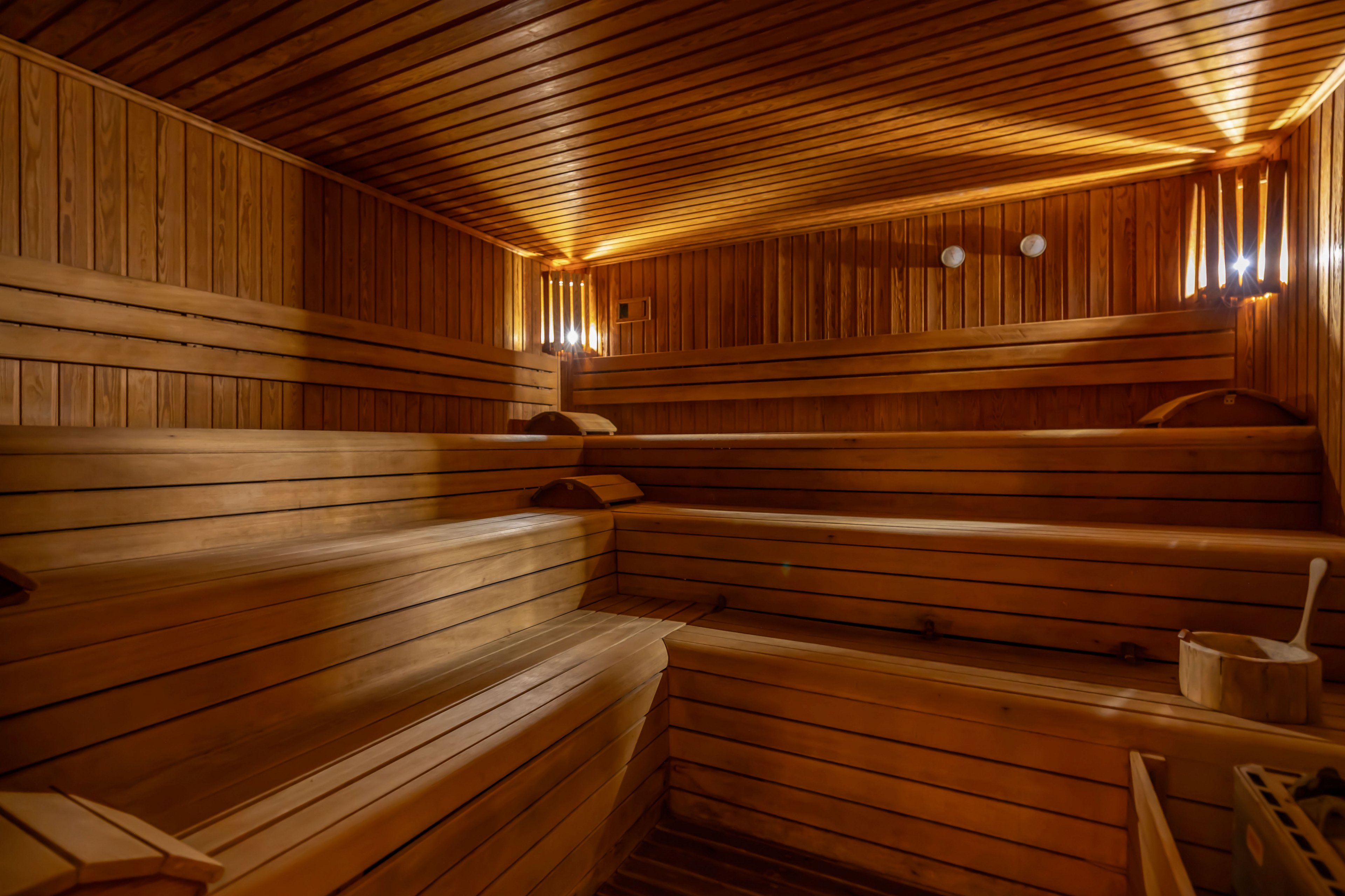 Image of an empty wooden sauna room with tiered benches. The space is lined with horizontal and vertical wooden panels, creating a uniform warm appearance. Soft wall lighting illuminates the interior, highlighting the texture of the wood. A wooden bucket and ladle are visible in the corner, emphasizing the traditional sauna setting.