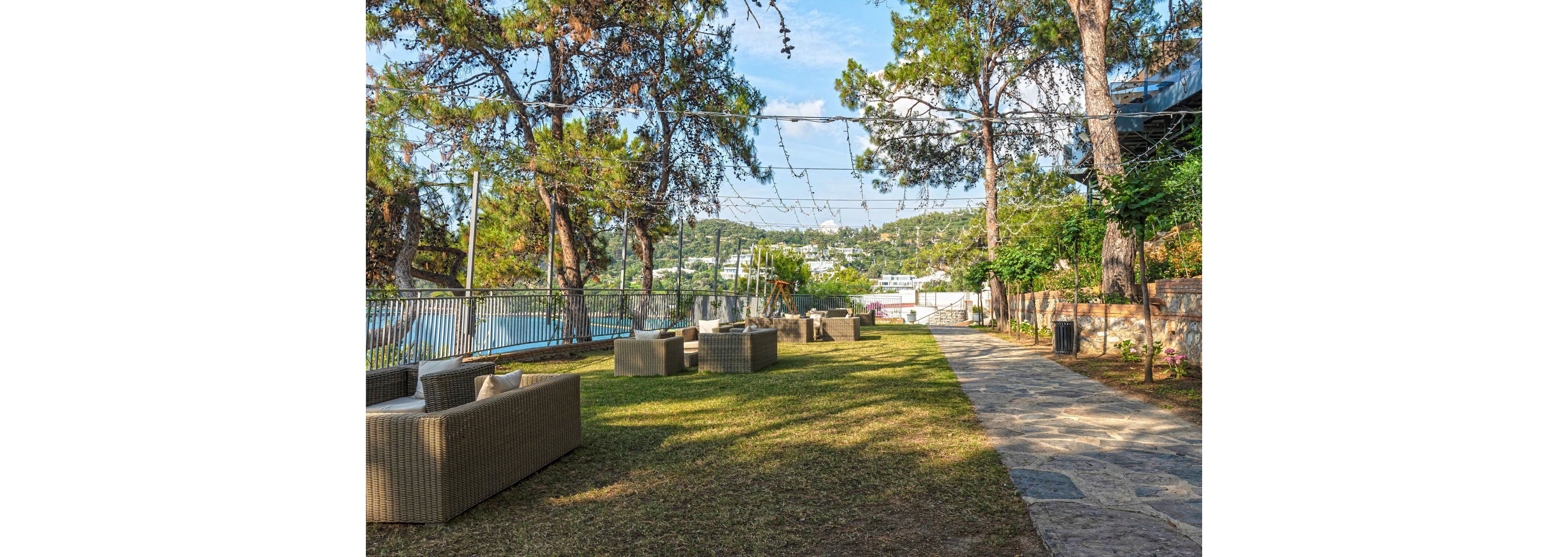 An outdoor lounge area features wicker sofas and armchairs arranged on a grassy lawn. Overhead string lights are suspended between tall trees, creating a relaxed atmosphere. A stone pathway runs alongside the seating area, bordered by a metal fence overlooking water. In the background, a hillside town and lush greenery are visible under a clear blue sky.