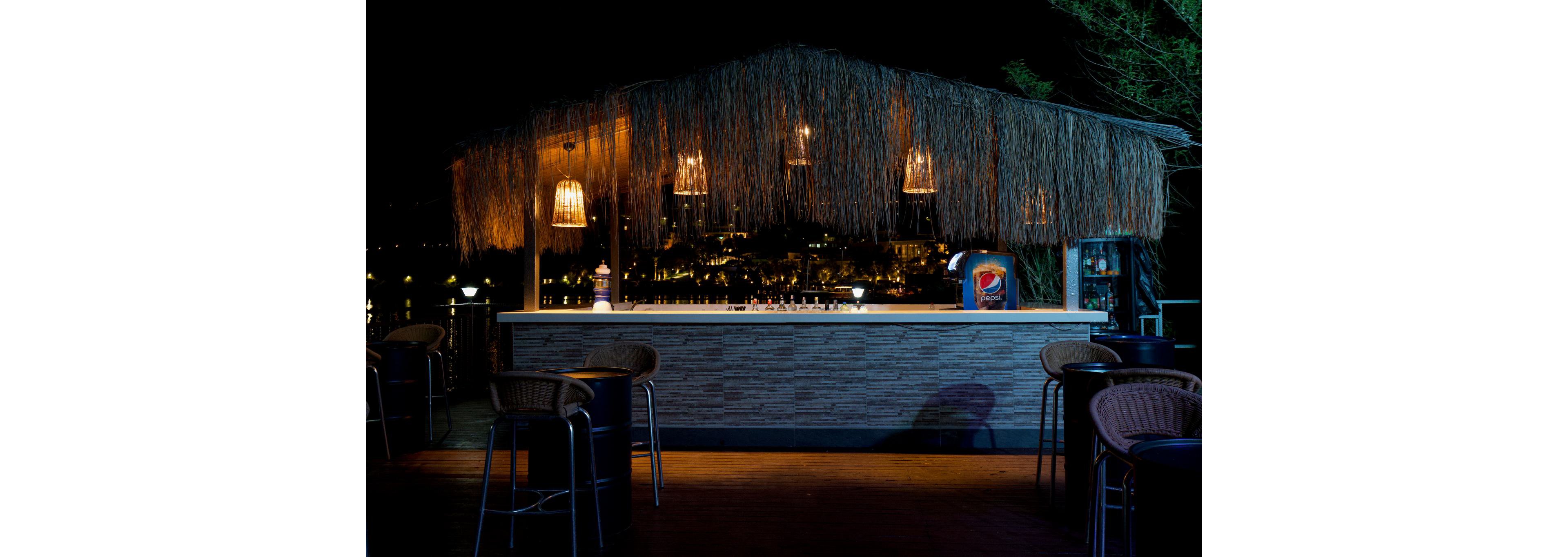 A thatched-roof outdoor bar is warmly lit against a dark night backdrop. Hanging pendant lights cast a soft glow over the counter, where bottles and bar accessories are arranged. High stools surround the bar, and a beverage cooler with a clearly visible Pepsi design stands to one side. The scene suggests a relaxed evening atmosphere in a resort or waterfront setting.