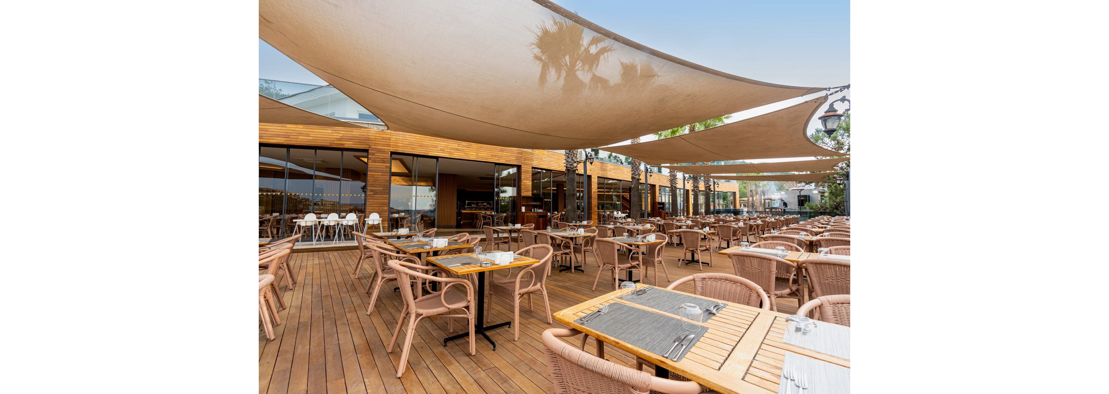An open-air restaurant terrace features numerous wooden tables and wicker chairs neatly arranged on a wooden deck. Large fabric shade sails stretch overhead, creating a comfortable, shaded dining environment. The background shows a contemporary building with large glass windows reflecting the surroundings. Table settings with placemats, glasses, and cutlery suggest the space is prepared for guests, while the overall scene appears calm and inviting.