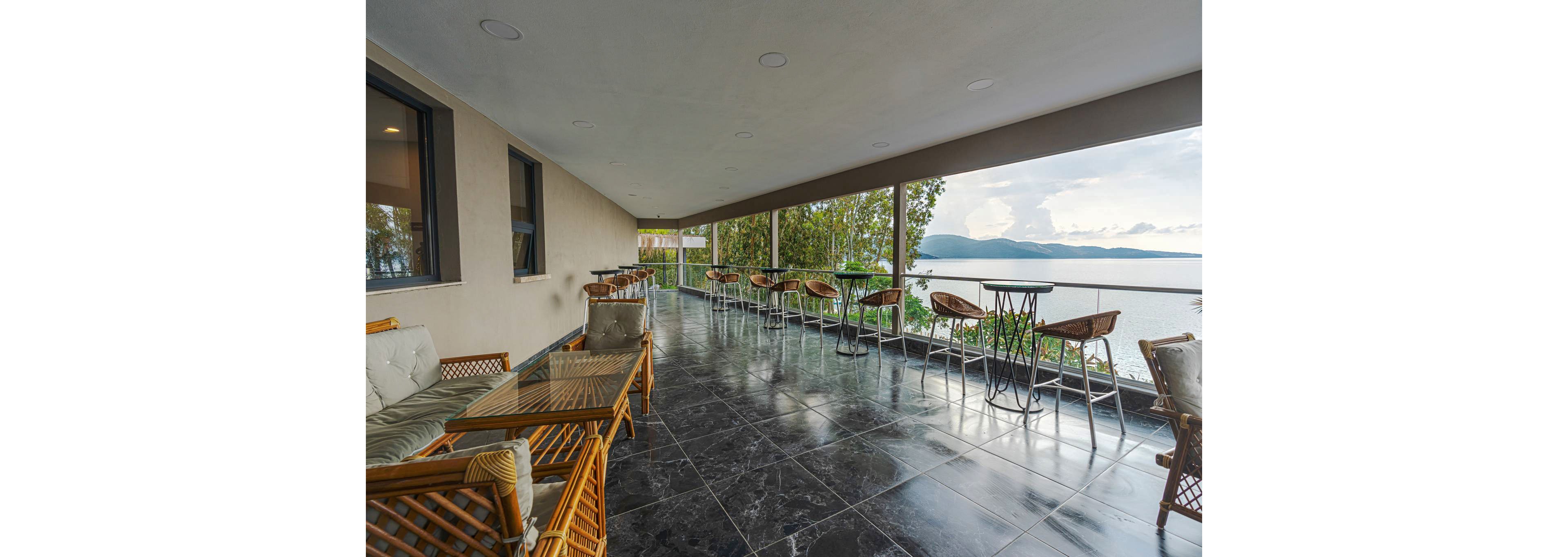 A spacious covered terrace overlooks a calm lake and distant mountains. Wicker chairs and glass-topped tables are arranged along the edge, creating a relaxed outdoor seating area. The floor is finished with glossy dark tiles that reflect the natural light. Large windows and open railings frame the scenic water and sky, suggesting a tranquil, contemporary hospitality setting.
