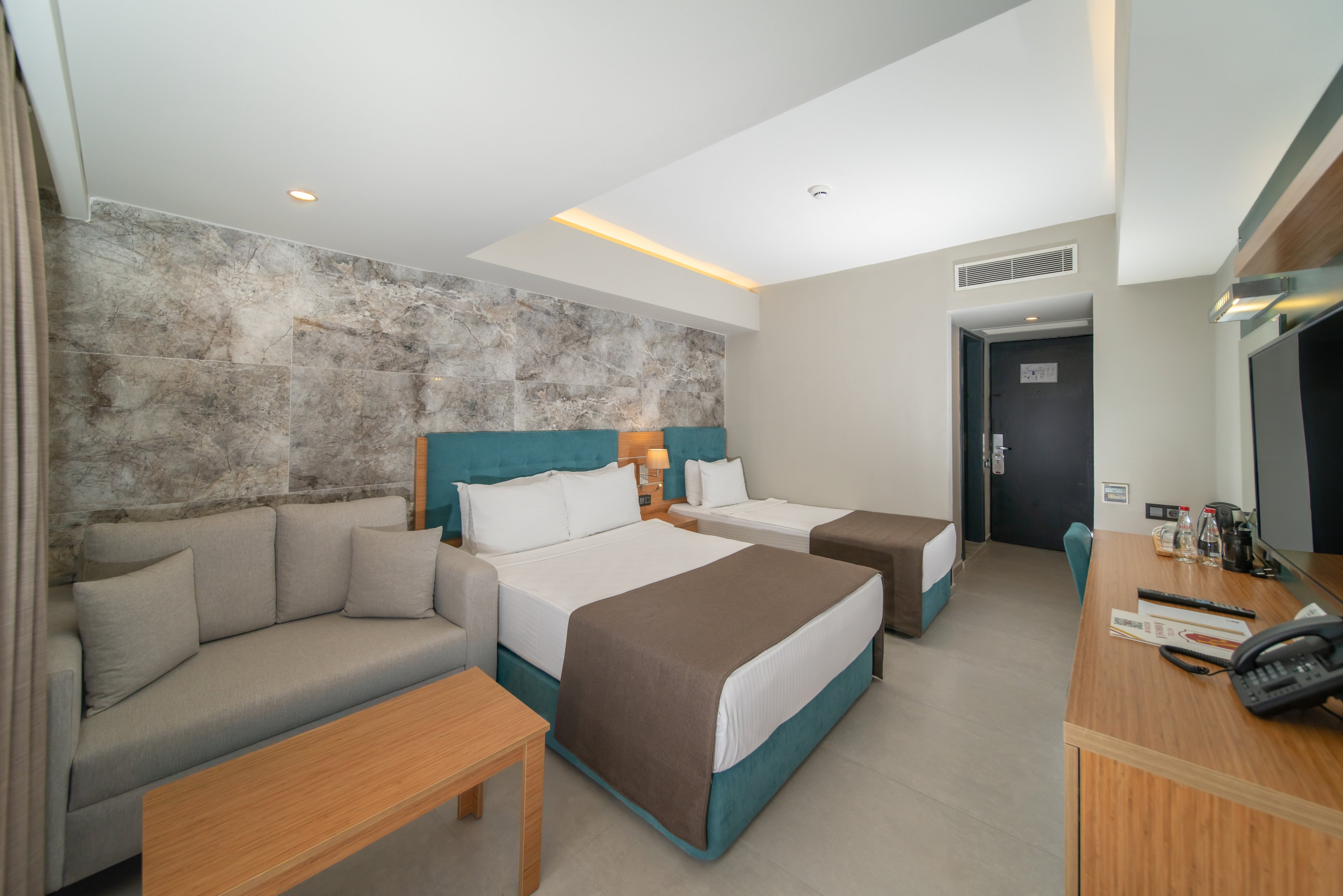 Contemporary hotel room interior featuring two neatly made twin beds with brown runners and teal bases. A light gray sofa and wooden coffee table sit beside a textured stone-look accent wall. Opposite the beds, a wooden desk holds a telephone, notepad, and various items, with a flat-screen TV mounted above. The space is bright and minimal, with neutral colors and clean lines creating a calm, welcoming atmosphere.
