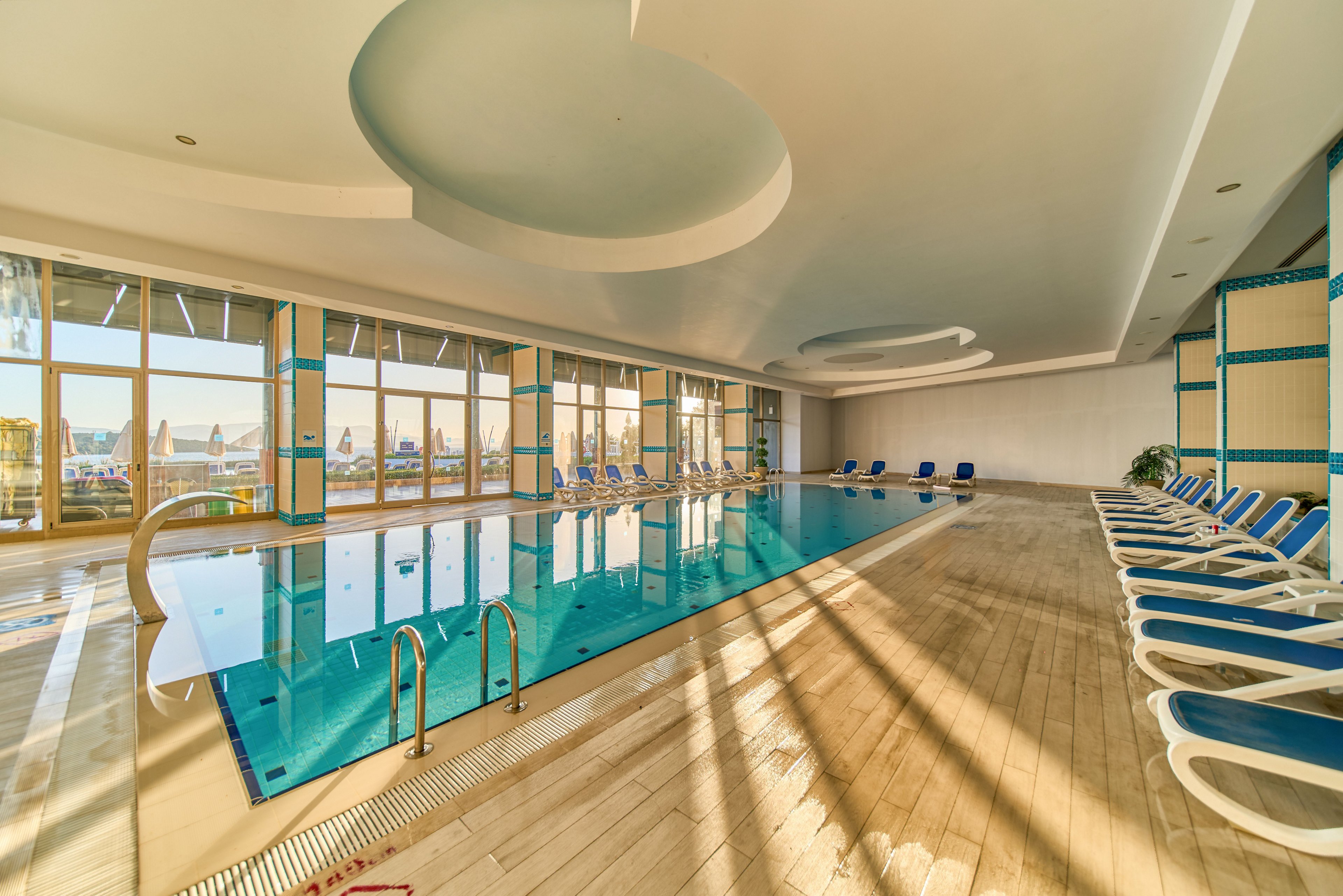 A bright indoor swimming pool is surrounded by rows of blue and white lounge chairs. Large floor-to-ceiling windows let in abundant sunlight, casting long shadows across the wooden deck. The calm water reflects the ceiling and window frames, creating a serene atmosphere. The space appears clean, modern, and ready for relaxation or exercise.