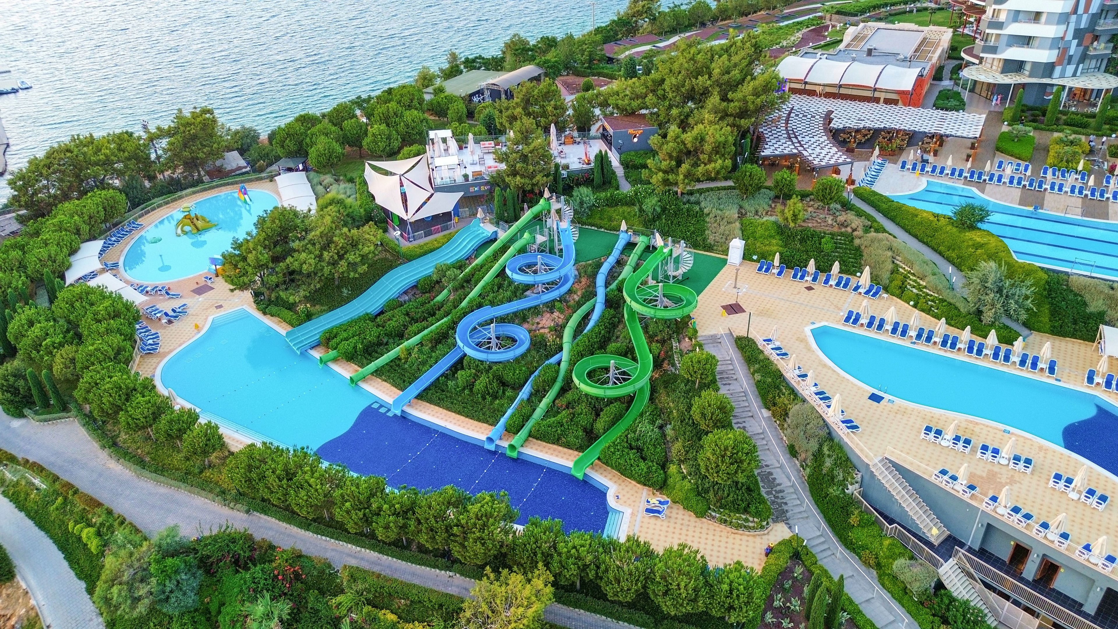 A large seaside resort water park is captured from above, showing multiple pools and winding water slides surrounded by lush greenery. Several bright blue slides twist down a landscaped hill into a main pool, with sun loungers neatly arranged on spacious decks. Additional pools, pathways, and buildings are visible, creating a vibrant vacation atmosphere next to the calm sea. The scene suggests a family-friendly destination focused on leisure, fun, and relaxation by the water.