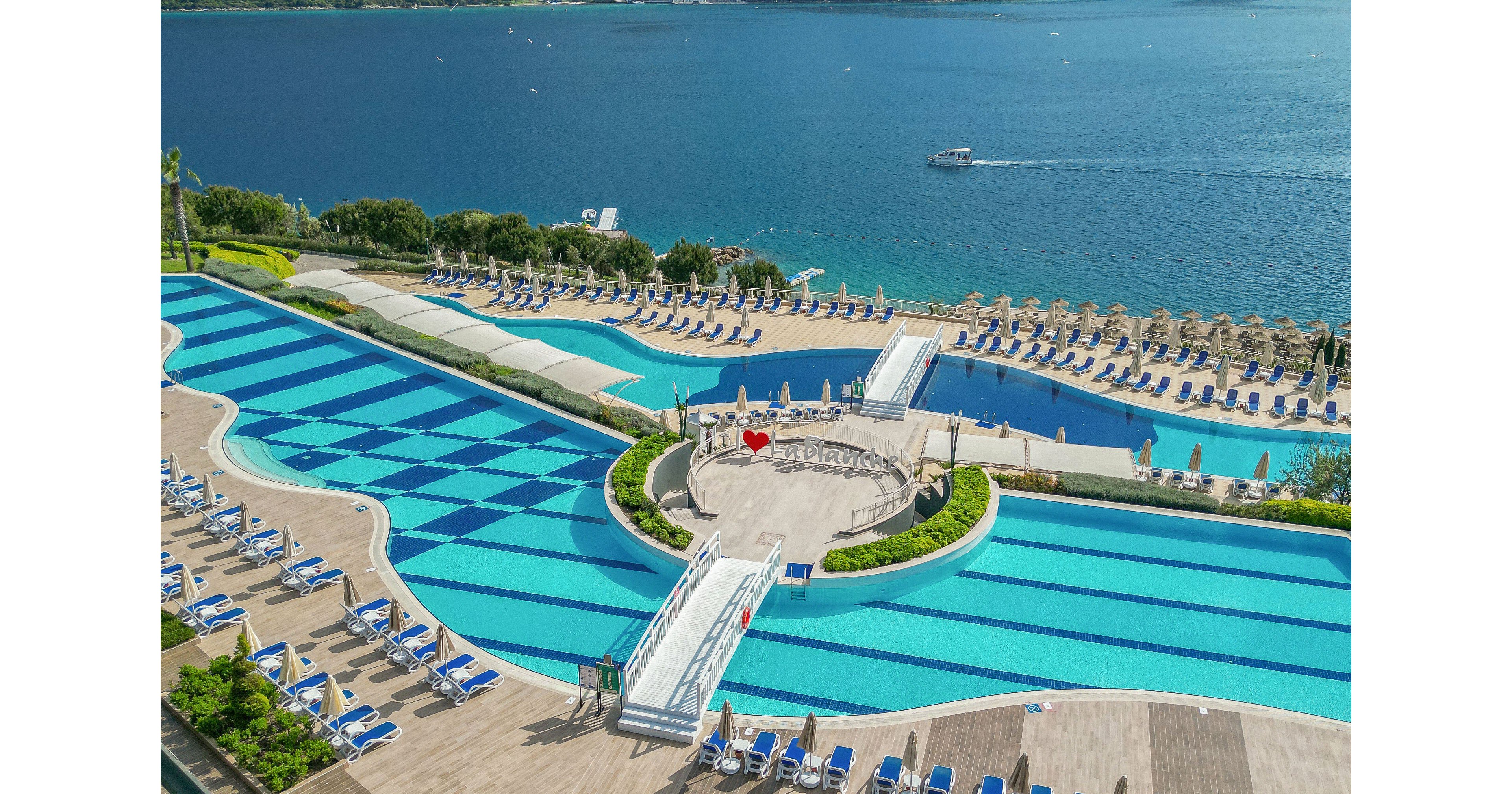 A large seaside resort features multiple expansive swimming pools arranged in geometric patterns. Rows of blue sun loungers and umbrellas line the pool decks, facing the water for relaxation and sunbathing. A central circular terrace with greenery and a white walkway connects different pool areas. Beyond the pools, a calm blue sea with a small boat and distant shoreline completes the scenic vacation setting.
